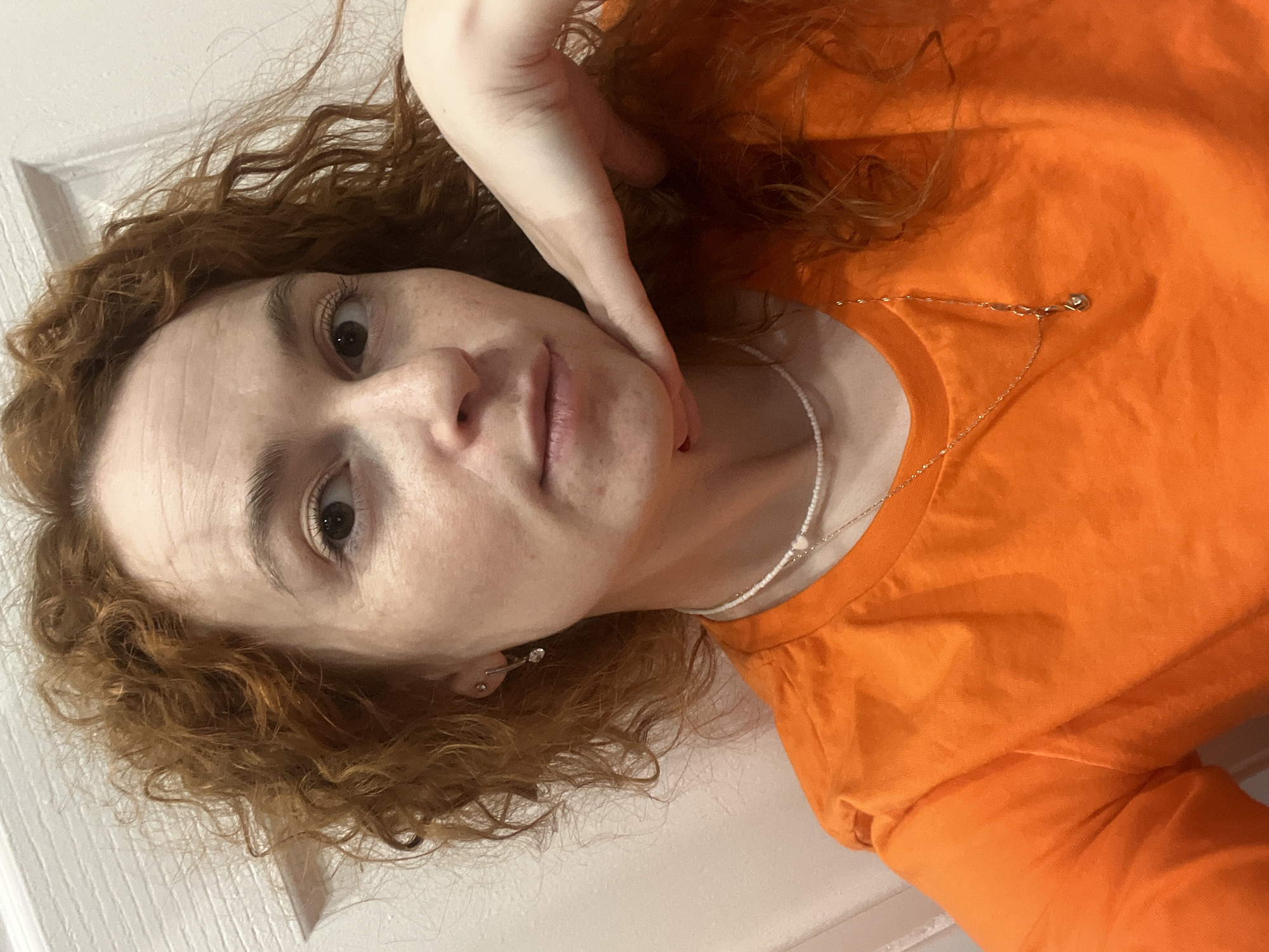A selfie shows a young woman with curly red hair wearing an orange T-shirt. She has fair skin and freckles. Her left hand is raised near her face, with her fingers lightly touching her cheek. She is wearing a delicate, layered necklace. A white door is partially visible in the upper left of the frame. It is impossible to determine if the picture was taken in the city of Chaikovsky, Russia based on the provided information.