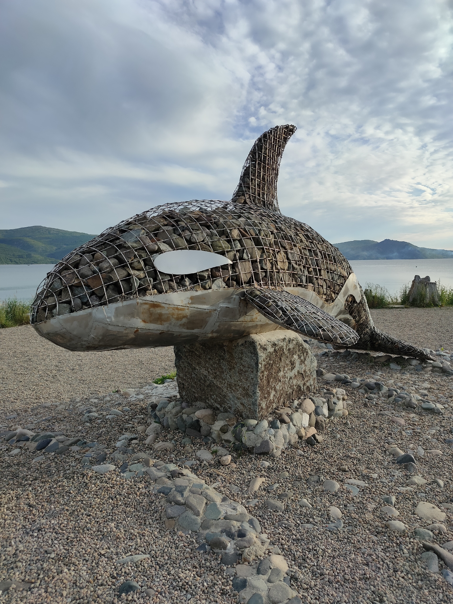 An outdoor, low-angle, full shot captures a large, stylized sculpture of an orca whale. The sculpture, crafted from a metal frame filled with stones, rests on a large, rough-hewn stone block. It is situated on a ground covered in small pebbles and larger stones. In the background, a calm body of water is visible, bordered by rolling hills covered in greenery. The sky above is cloudy with patches of blue peeking through. The sculpture is located at the Magadan Bay waterfront in Magadan, Russia. There are no people visible in the frame.