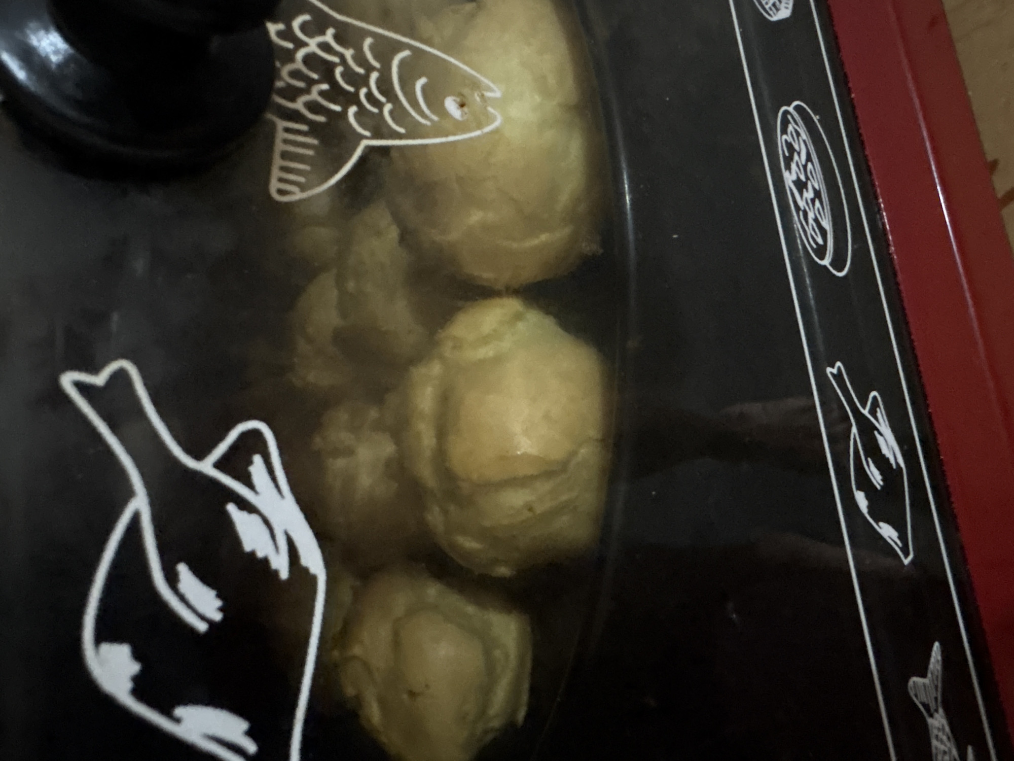 The image shows a close-up view of a red and black appliance, likely a small oven or grill, with a glass lid. Inside the appliance, several golden-brown, round food items are visible, possibly pastries or baked goods. The glass lid has white line drawings on it, including a stylized fish on the left and a bird-like figure on the right, with more decorative circular patterns in between. The appliance appears to be in a domestic setting, with hints of a wooden surface or floor visible in the background. The lighting suggests it is daytime, but no specific time of day can be determined. There is no visible text in the image.