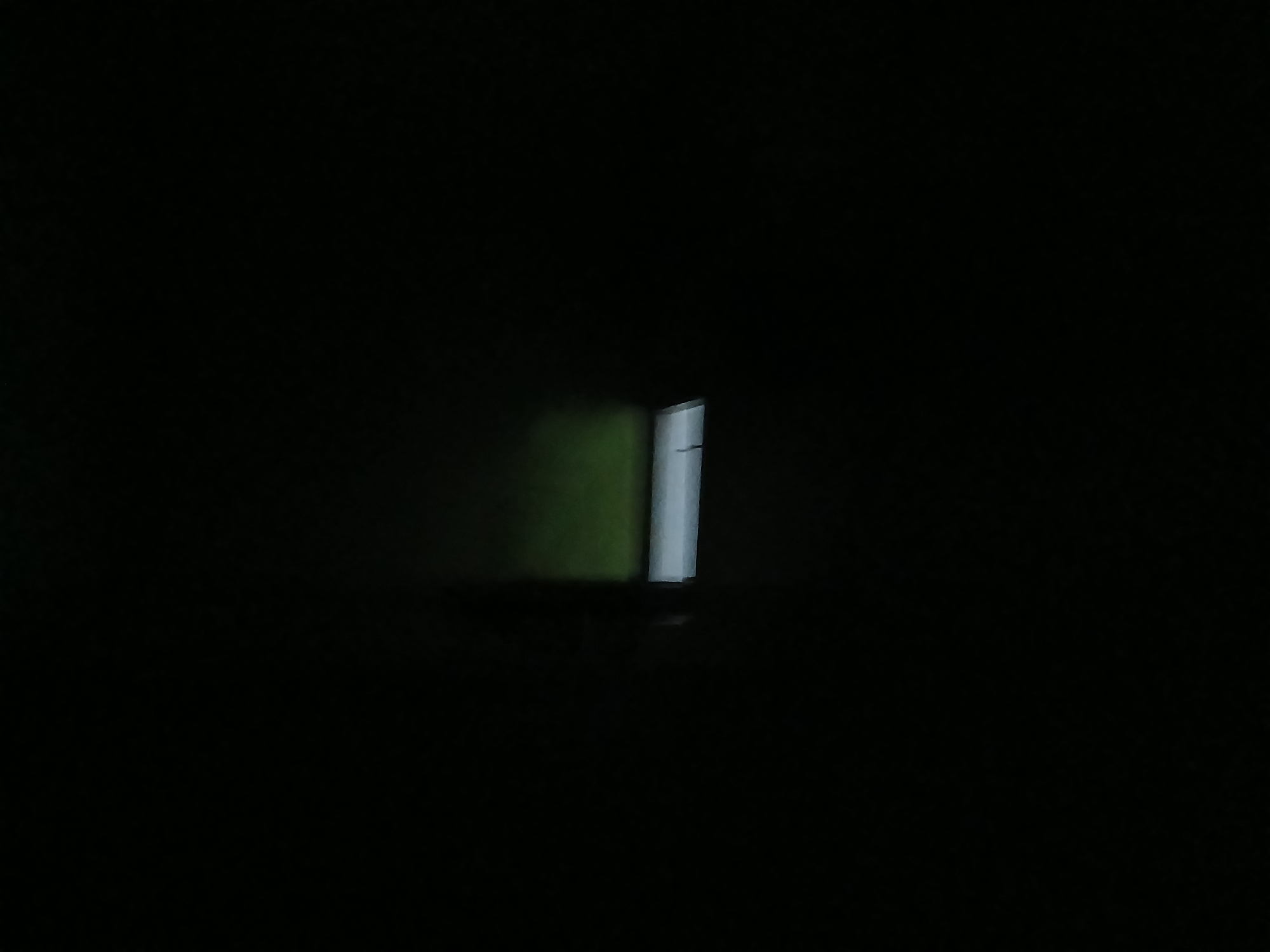 In a dimly lit environment, a rectangular light fixture is visible horizontally across the center of the frame. Above the light, a faint green hue suggests a wall or ceiling illuminated by an unseen light source. The surrounding area is predominantly black, indicating deep shadow or darkness. The image appears to be taken indoors, possibly in a room or hallway, in Tenggarang, Indonesia, although specific details of the location are not discernible from the visual information provided. There are no people, actions, or interactions depicted in the scene.