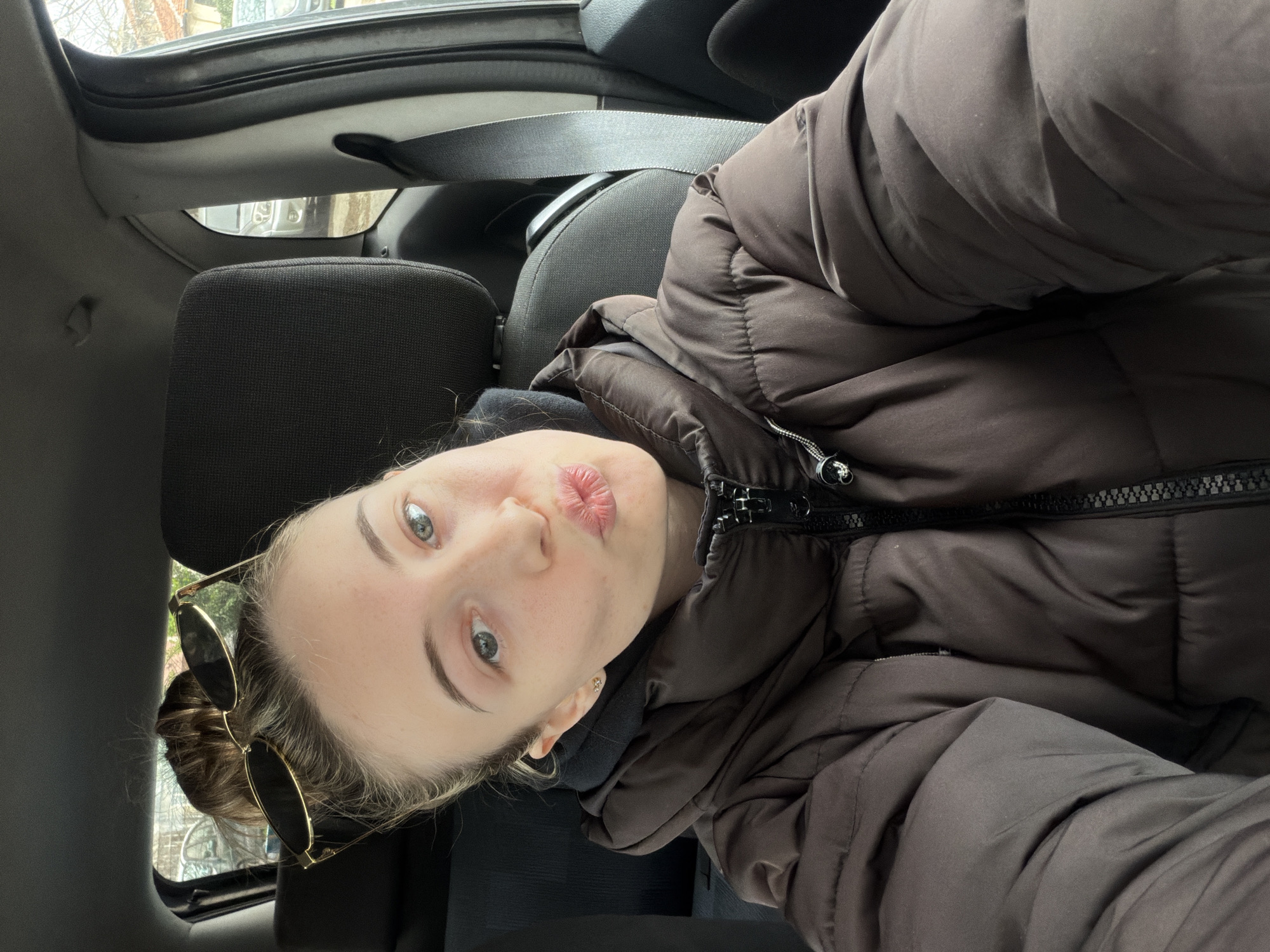 The image shows a person in the passenger seat of a car. The person is wearing a brown puffy jacket and has sunglasses tucked into their hair. The car appears to be parked, with the interior visible, including the seats and safety belt. The location is not discernible in the image. The person is puckering their lips. The environment includes car interior elements like seats, headrests, seatbelts, and interior ceiling. Without additional information, the city of Plovdiv, Bulgaria cannot be confirmed in the description.