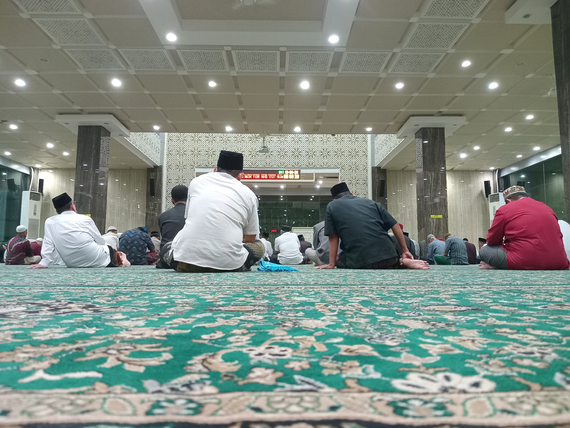 The image depicts the interior of a mosque in Randuagung, Indonesia, where a group of men are seated on a green carpet with an intricate pattern. They appear to be engaged in prayer or attending a religious service. The perspective is from a low angle, looking across the carpet towards the back of the seated individuals. The mosque features a modern ceiling with recessed lighting and decorative panels. Pillars with a dark, patterned finish are visible, as is a large digital display on a wall in the background, likely showing prayer times. The overall environment suggests a clean, well-maintained, and possibly spacious place of worship.
