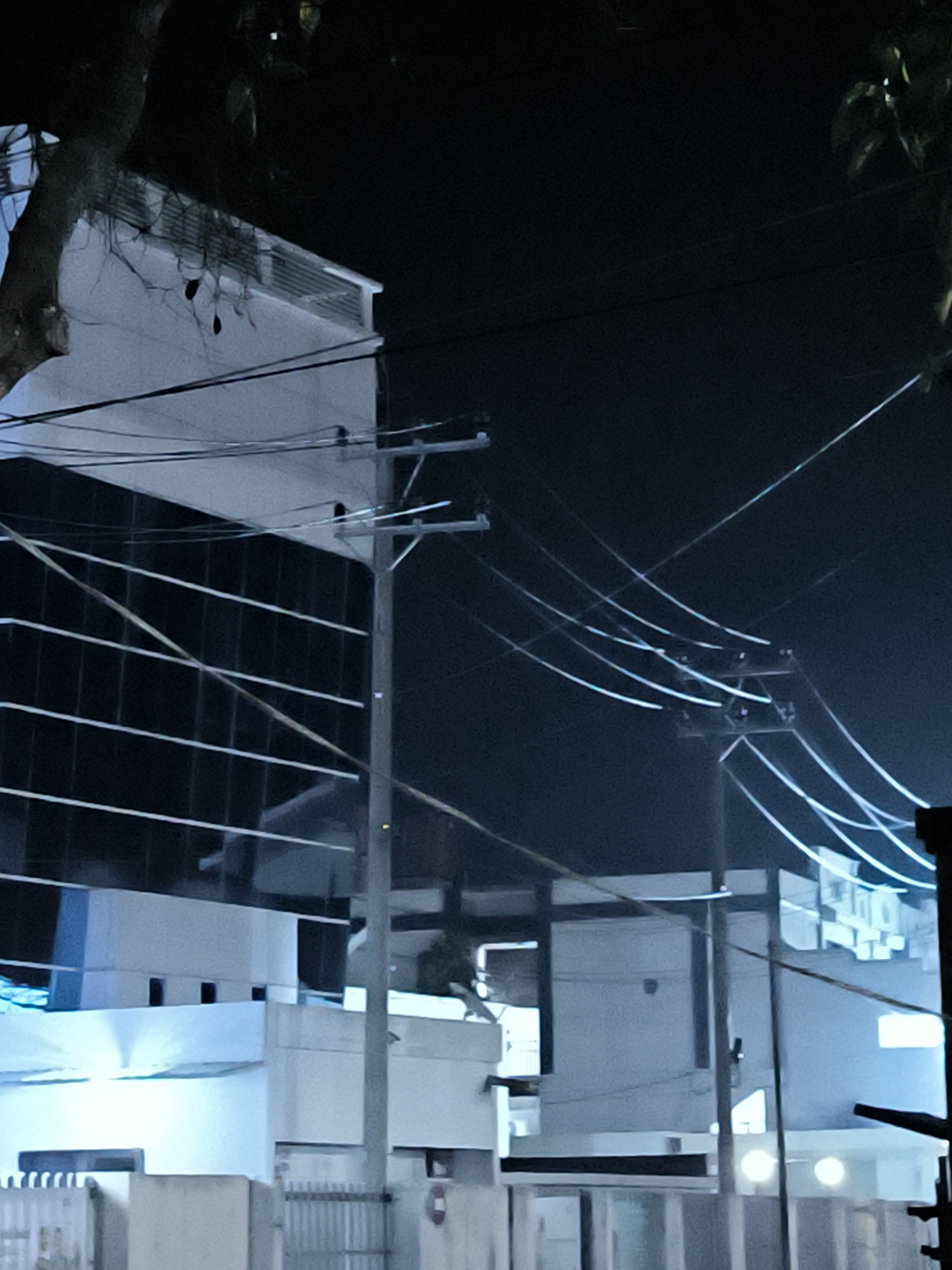 The image shows a nighttime scene in Lhokseumawe, Indonesia, featuring a mix of residential and commercial buildings. Power lines stretch across the sky, supported by concrete utility poles. The building on the left appears to be a modern, multi-story structure with dark, reflective windows. Lower buildings with white exteriors are visible in the foreground and middle ground, with possible windows and doors. Trees are partially visible in the frame's periphery. A fence or gate can be seen in the immediate foreground. The overall lighting is dim, with the buildings and power lines reflecting ambient light.