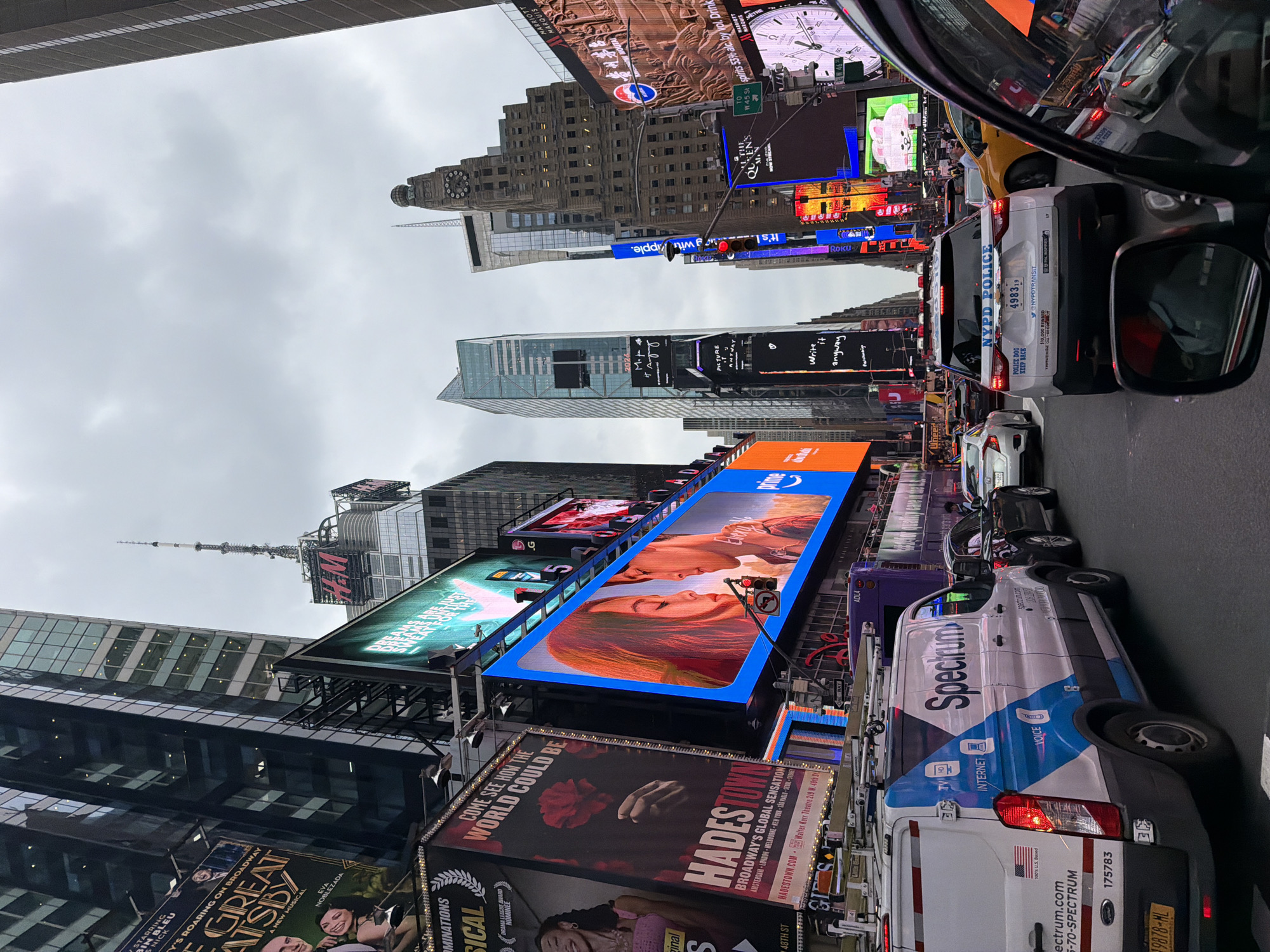 The image depicts a street scene in Times Square, New York City, USA, under an overcast sky. The view is from within a vehicle, looking forward at traffic and the surrounding buildings adorned with numerous large digital billboards.
In the foreground, the right side of a dark-colored SUV is visible, including its side mirror which reflects the surrounding environment. Ahead, a white NYPD police car with flashing lights is prominent in the lane, displaying the text "NYPD POLICE" and license plate "4983 19". To the left of the police car is a white van with "Spectrum" branding and logos for TV, HD, and Internet. Further ahead, several other vehicles are stopped in traffic, including a white sedan and a yellow taxi.
The background is dominated by towering buildings with a dense concentration of illuminated billboards displaying advertisements for various products and entertainment, including a prominent blue screen showing a couple, a large billboard for "The Great Gatsby" musical, and an H&M advertisement. The overall atmosphere is urban and commercial, typical of Times Square. People are visible in some of the vehicles and on the sidewalks, though their actions are not clearly discernible. The street is paved, and traffic signals are present.