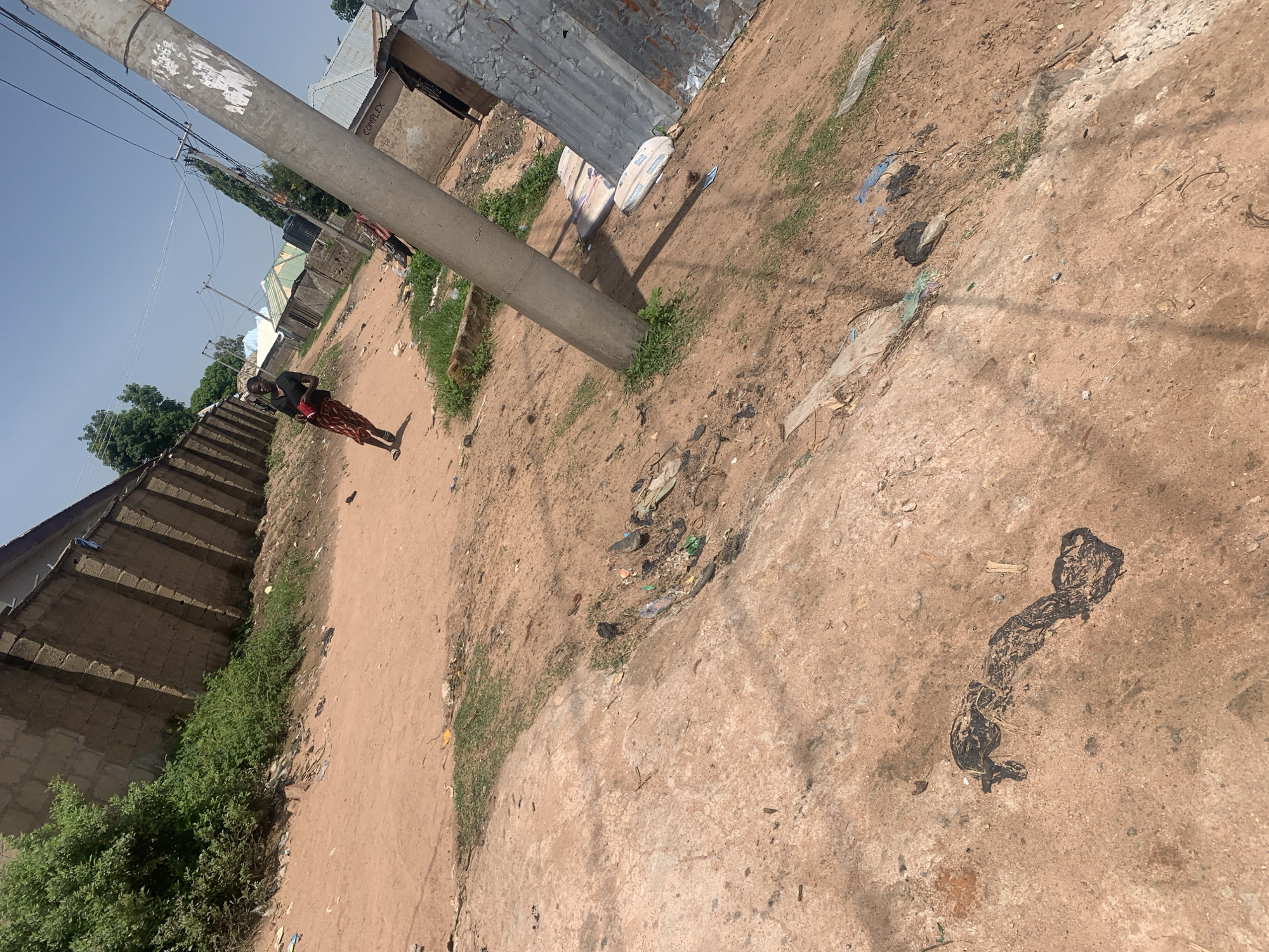In Jauro Yinu, Nigeria, a dirt road winds through a residential area characterized by buildings with concrete block walls and corrugated metal roofs. A young woman, dressed in a black top and a patterned skirt, walks down the road, holding a red object in her left hand. In the foreground, a concrete pole stands next to a cluster of trash and some green grass. Further down the road, additional buildings and power lines are visible under a clear blue sky. The ground is dry and dusty, with scattered debris.