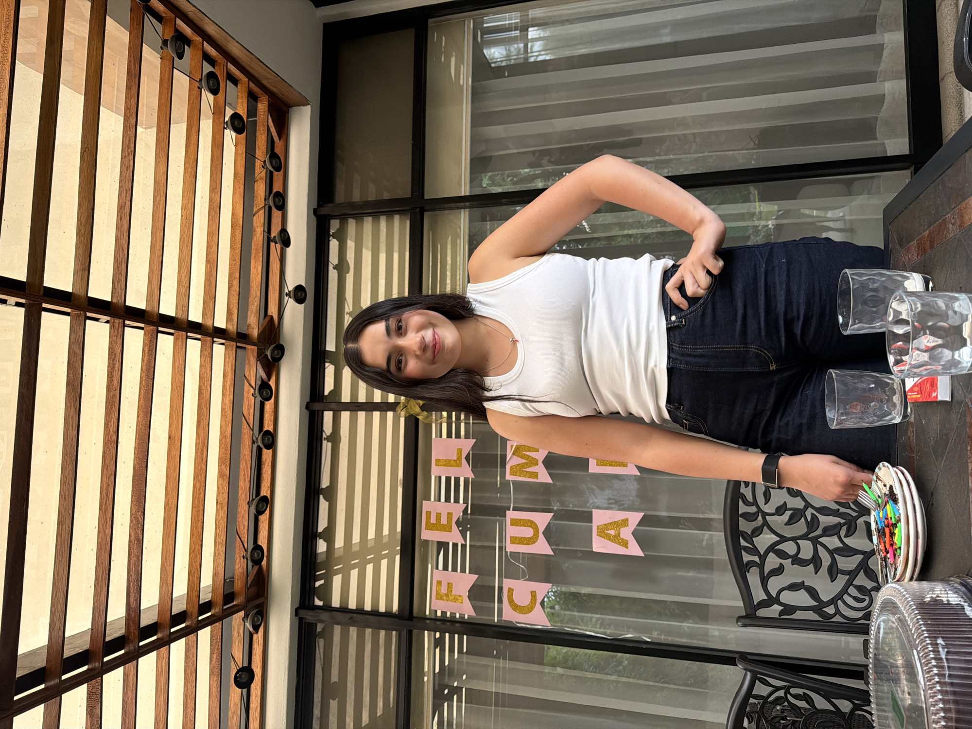 The image shows a woman standing at a table on a patio or balcony. She is wearing a white tank top and dark jeans. She is smiling and looking at the camera. On the table, there is a decorated cake, glasses, and other items. In the background, there is a window with a "FELIZ CUMPLE" banner hanging on it. The patio is enclosed by a wooden structure with lights. The location is Barrio de Nuevo León, Mexico.