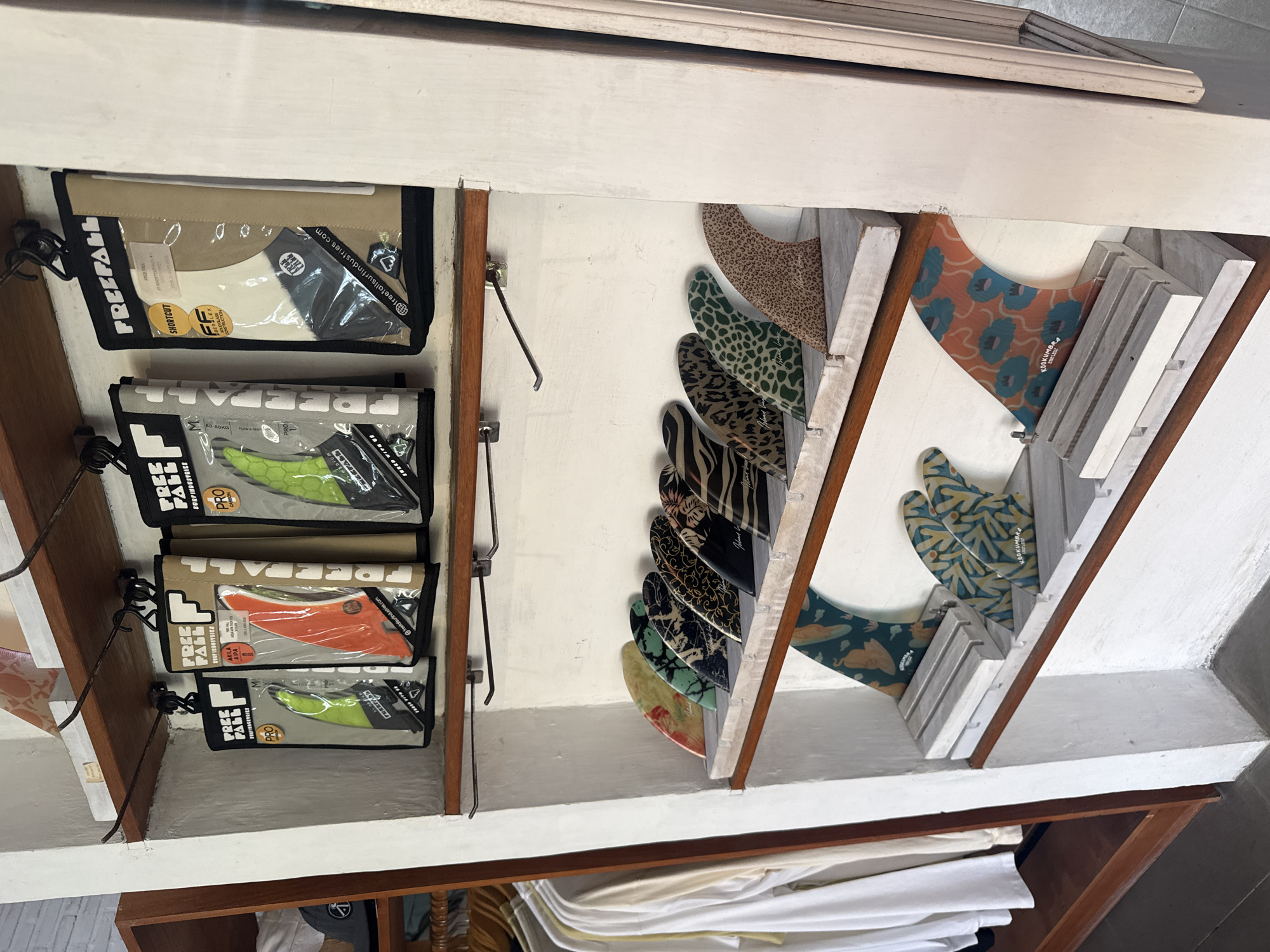 The photograph depicts an interior scene, presumably a shop or retail space, presenting a display of surfboard fins and their packaging. On the left side, there are several black pouches with transparent windows, labeled with the brand "FREE FALL" and other product information. The pouches are mounted on a rack using metal hooks. Visible inside the pouches are the fins, showcasing different colors such as white, green, and orange, and other technical details about the fins.
To the right, a collection of surfboard fins is organized within a wooden display structure. The fins are arranged vertically, exhibiting a variety of patterns and colors, including leopard print, zebra stripes, floral motifs, and stylized graphic designs. The fins are marked with branding, one visible instance reading "KOOKUMBRA".
The overall environment appears to be a retail space, with a neutral, off-white background. There are articles of clothing stacked on the shelf below. The scene suggests a business focused on surfing equipment and related accessories, located in an unspecified urban area.