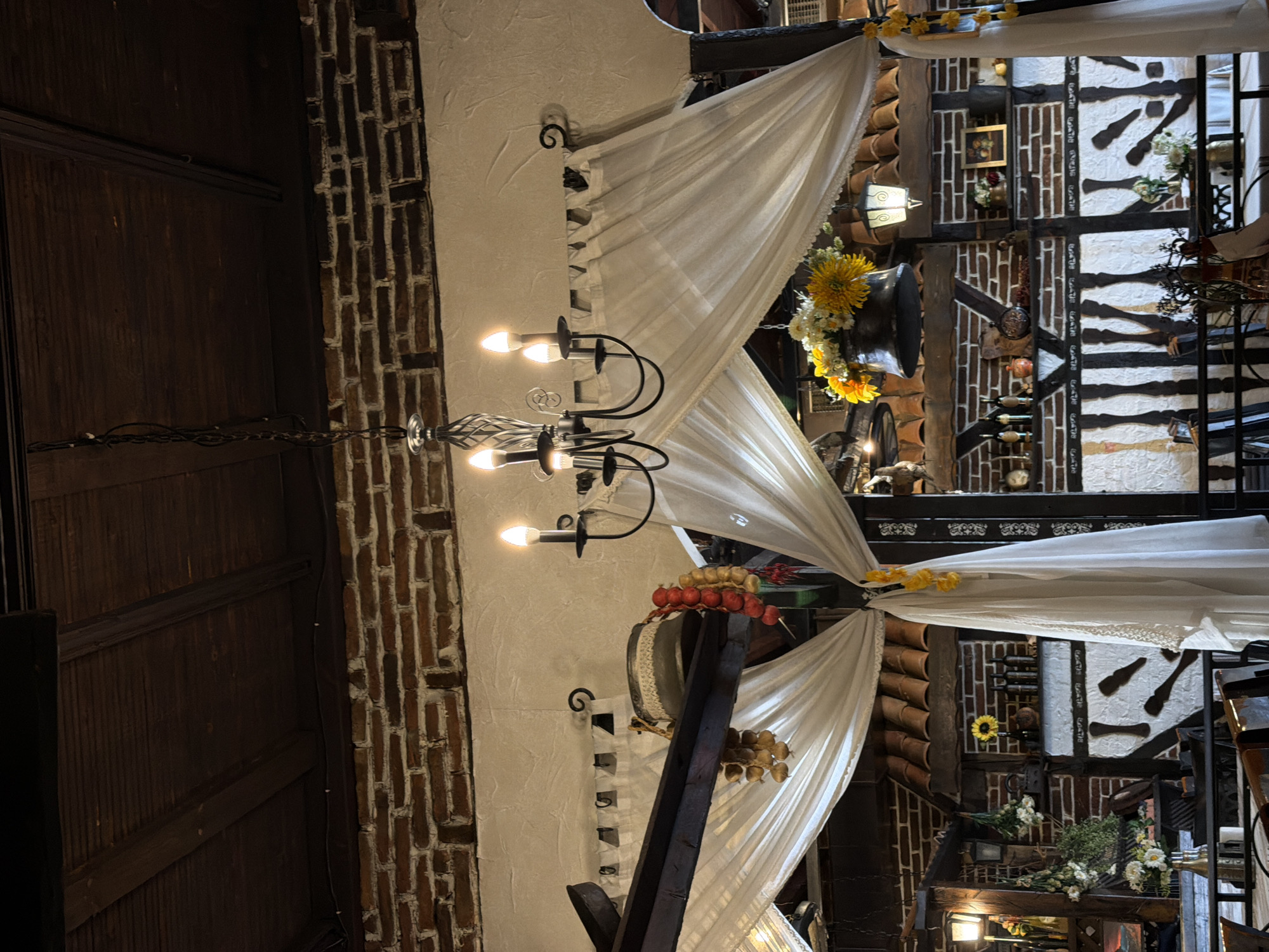 The image depicts the interior of a building with decorative elements, possibly a restaurant or event space. It features a chandelier with multiple lights, positioned near a white draped fabric ceiling adorned with yellow floral arrangements. The building's interior showcases exposed brickwork, white plaster walls, and dark wooden beams, suggesting a rustic or traditional style. There are decorative items hanging on the walls, and the presence of flowers and other ornaments gives the scene a festive or celebratory atmosphere. Based on the decorative style and architecture, the location is likely in Varna, Bulgaria, where similar architectural and decorative styles are common.