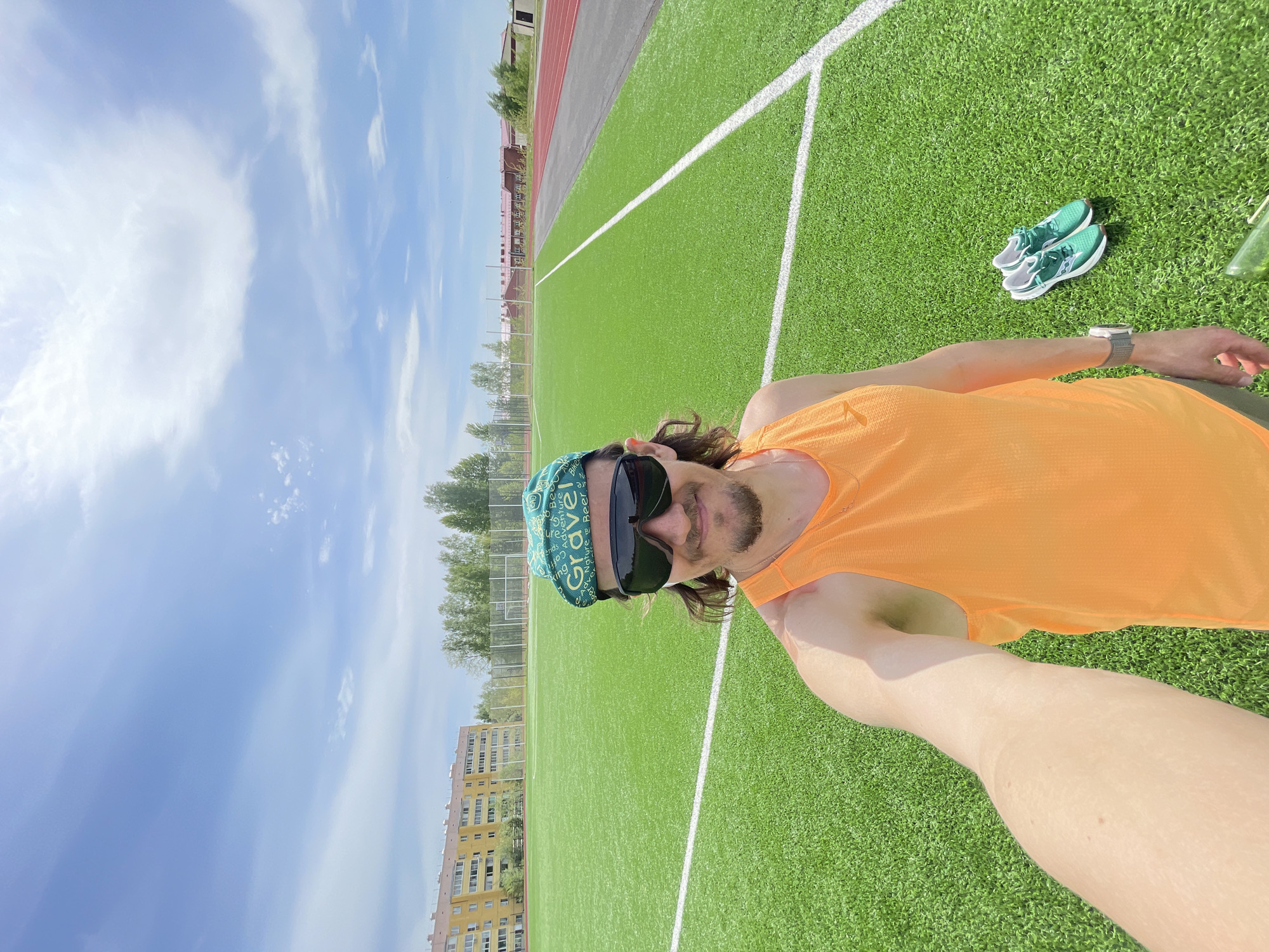 A selfie was taken on a sunny day at a green sports field, likely a football or soccer field, with white lines marking the playing area. The subject, a person wearing an orange tank top, sunglasses, and a green headscarf with white lettering (possibly "Gravel"), is positioned in the foreground. A pair of teal and white sneakers are placed to the right of the subject. A red running track is visible in the background. The location is possibly Noviy Urengoy, Russia, based on the architectural style of the buildings in the distance.