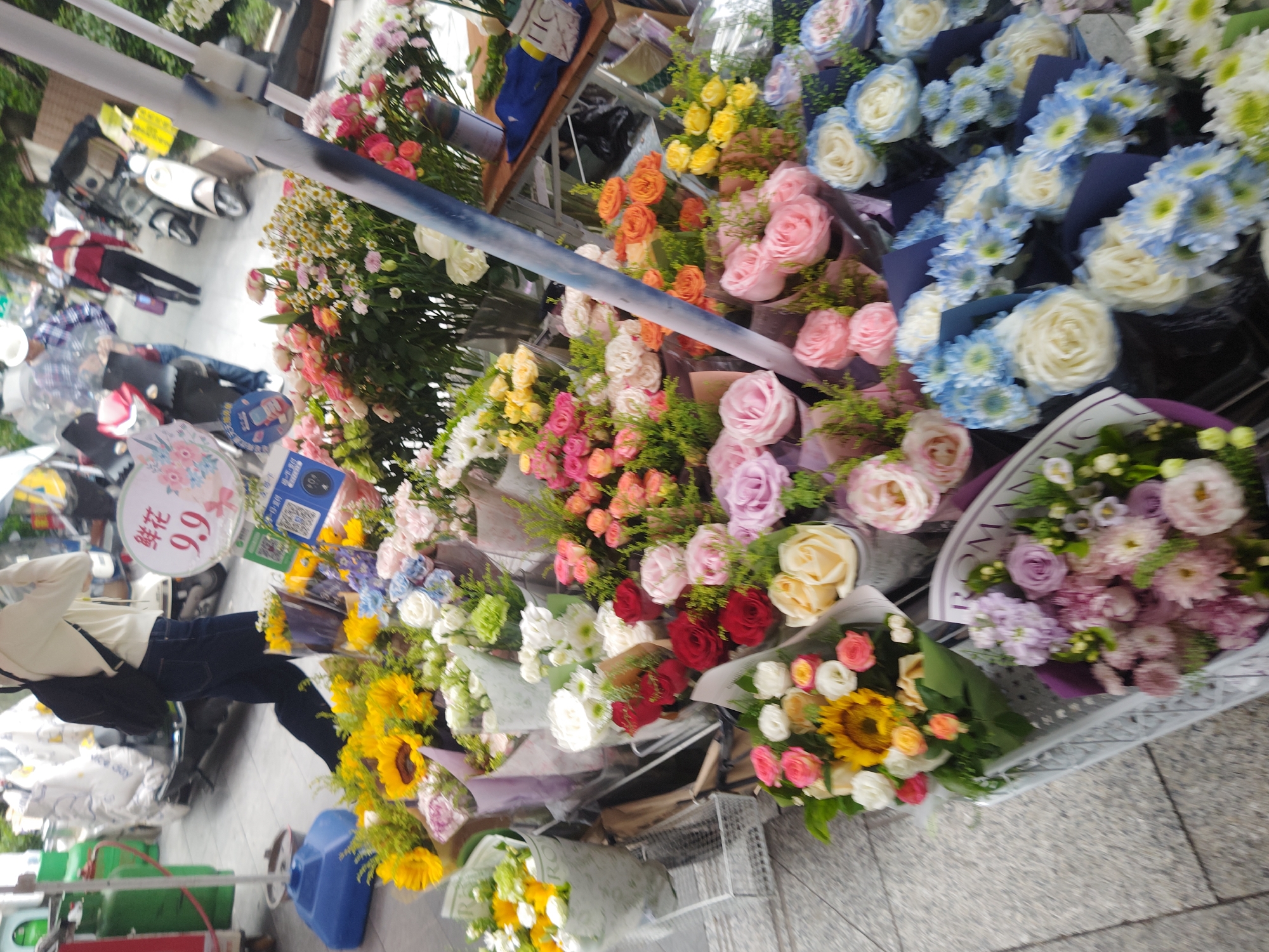 The image shows an outdoor flower stall in Chongqing High-tech Zone, China. A variety of flower bouquets, including roses, sunflowers, and daisies, are on display. A person wearing a beige top and blue jeans stands beside the stall. In the background, there are scooters and pedestrians. A sign with the price "9.9" is visible.