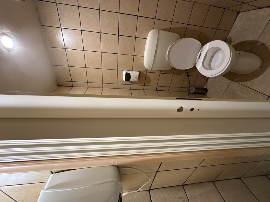 This image captures a brightly lit bathroom or public restroom interior, taken with a perspective that is rotated 90 degrees counter-clockwise. Light beige square tiles with dark grout lines cover both the walls and floor, appearing to extend from the top to the bottom of the frame.
A white ceramic toilet with its lid and seat open is positioned in the upper-right section of the rotated image. To its left, a white hand sanitizer dispenser with a dark accent is mounted on the tiled wall. Below the toilet, a metallic cylindrical object, likely a toilet brush holder, is visible near the floor.
A recessed circular light fixture illuminates the upper-left portion of the frame, casting a bright glow. A long, white, rectangular fixture, possibly a vanity or shelf, spans horizontally across the middle, featuring two circular cutouts and a smaller hole. In the immediate foreground, another white rectangular structure, potentially a baseboard or part of a larger fixture, runs along the bottom, with a white electrical cord snaking across the tiled surface.
The tiles and grout show some signs of use and minor discoloration. There are no people present, and no specific activity is taking place. The scene is static and well-lit by artificial light, suggesting an indoor setting where natural light is not a primary factor. No visible text is discernible on any objects. The location is Nea Erithrea, Greece.