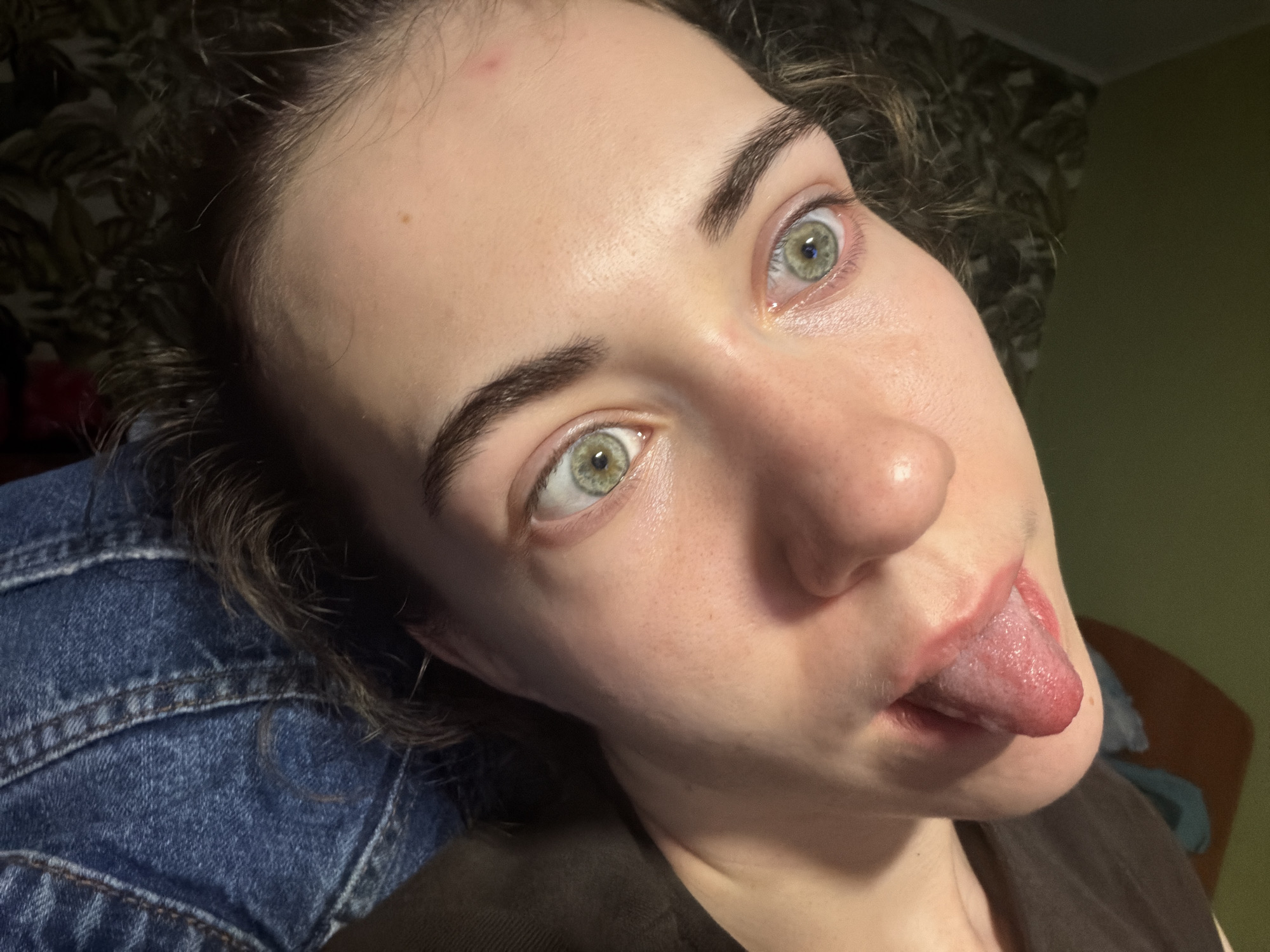 The image shows a close-up of a young person's face, with their eyes wide open and their tongue sticking out. They are wearing a dark brown shirt and what appears to be a denim jacket. The background is blurred, but it seems to be an indoor setting with some patterned wallpaper and a plain green wall. The lighting suggests it is evening or night. There is no visible information in the image that links it to the city of Zavodoukovsk, Russia.