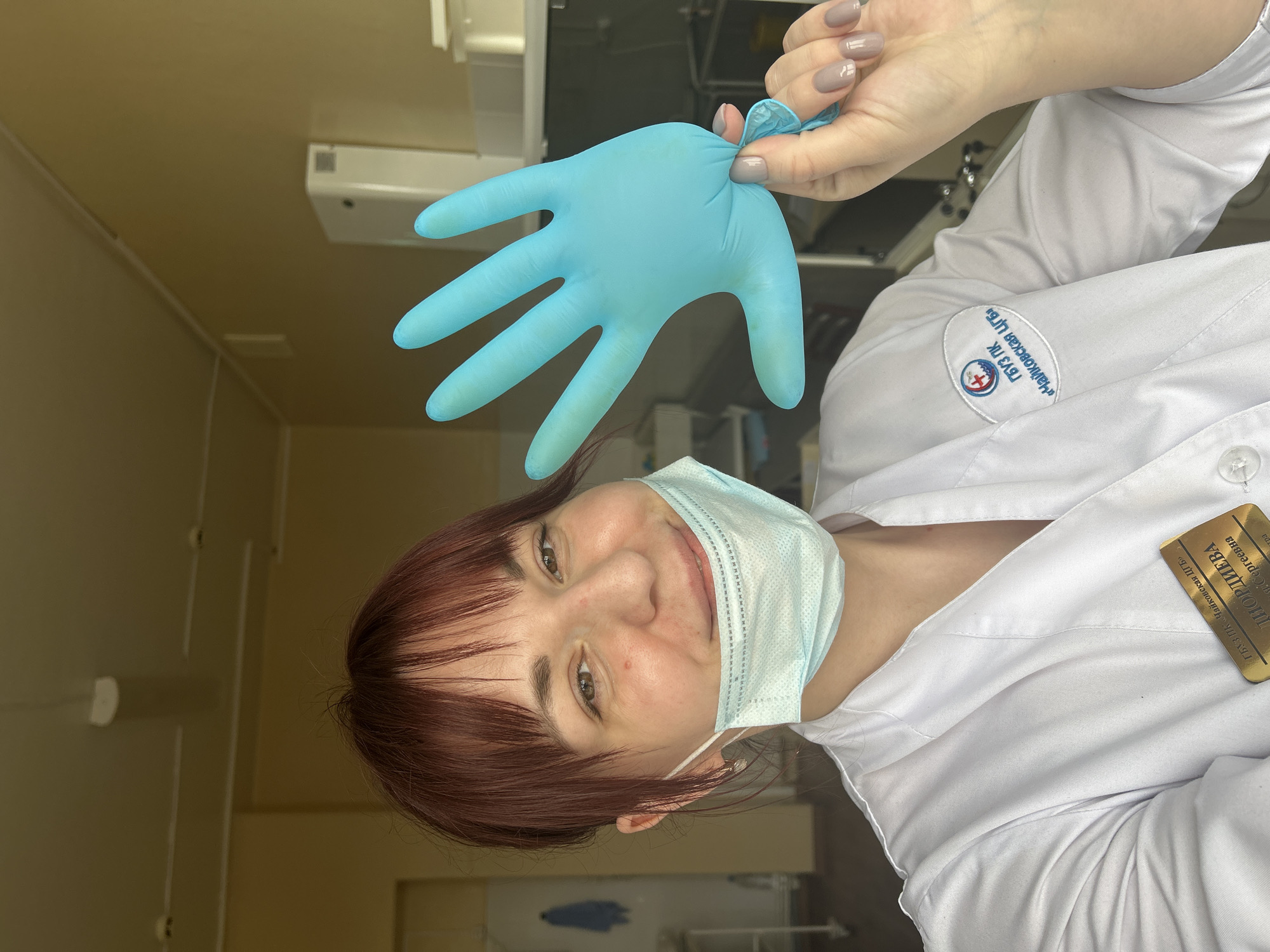 A female medical professional is photographed indoors. She is wearing a white lab coat, a light blue surgical mask, and is holding a light blue medical glove. Her brown hair is cut in a fringe style. She is smiling at the camera. A name badge on her coat reads "ИОРЯЕВА Полина Андреевна ГБУЗ ПК "Чайковская ЦГБ"." The background is an interior medical setting. This indicates that the photo was taken within the Chaikovskaya Central City Hospital, which is located in the city of Фоки, Russia.