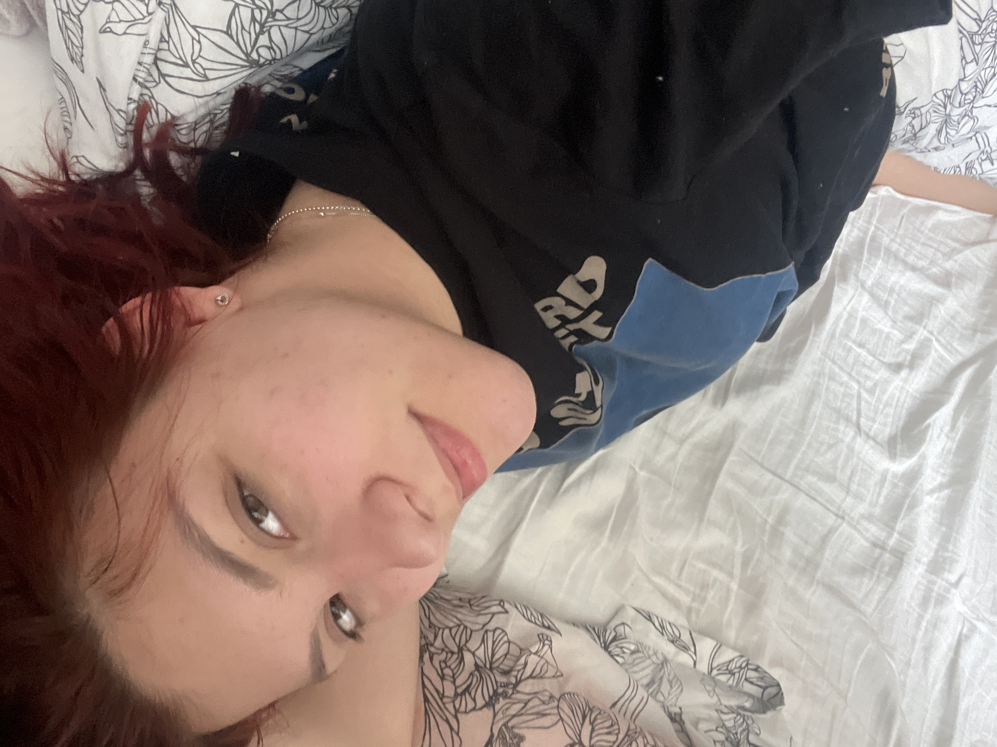 A young adult female is lying on a white bed with floral print sheets and pillowcases. She is angled toward the camera, her head resting slightly downward. She has reddish-brown hair, fair skin, and wears a black and blue hooded sweatshirt. A silver necklace is visible around her neck, and she appears to have an earring. The location cannot be determined to be in Sofia, Bulgaria based on the image alone.