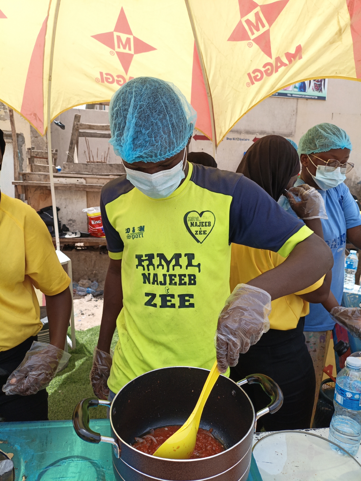 The image depicts an outdoor scene in Tarauni, Nigeria, where individuals are preparing food. A person in the foreground, wearing a blue hairnet and a light blue surgical mask, is stirring a pot of red sauce with a yellow ladle. They are also wearing a bright yellow and dark blue t-shirt with the text "HML NAJEEB & ZEE" and the "SPORT" logo. They are also wearing clear plastic gloves, as are the other individuals visible. To the left, another person in a yellow t-shirt is partially visible. In the background, under a yellow umbrella with a red star logo and the word "MAGGI" printed on it, other individuals can be seen. One person is wearing a black hijab and a blue t-shirt, while another to their right wears a light blue hairnet, glasses, a surgical mask, and a light blue t-shirt. Various items are visible in the background, including a white bucket with a red label, plastic bottles of water, and what appears to be a food stall setup. The environment suggests a busy marketplace or outdoor event.