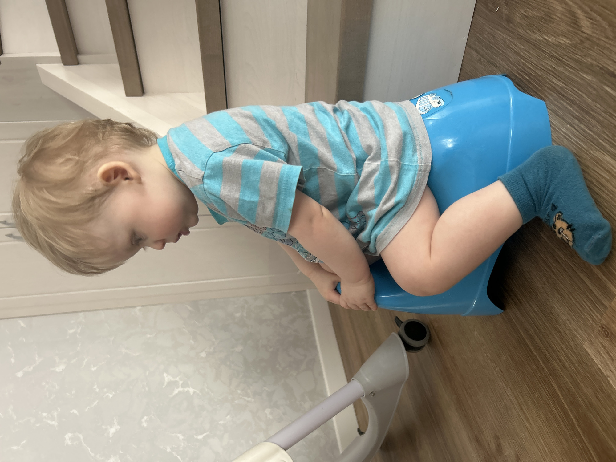 A young child wearing a blue and gray striped shirt and a blue sock with a bear design is sitting on a blue potty chair. The potty chair is on a wooden floor. The child is leaning forward with their head down and their hands on the potty chair. Stairs are visible in the background. Part of a walker or other support device is visible in the lower left corner of the image. The photograph was likely taken indoors. The location in the city of Chaikovsky, Russia, is not evident from the image.