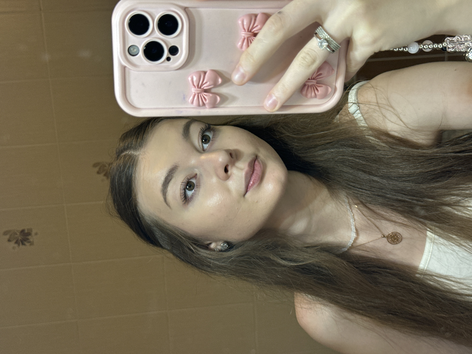 A young woman is taking a selfie in a bathroom. She has long brown hair and is wearing a white tank top, a necklace, and earrings. She is holding a pink phone case decorated with bows. The bathroom walls are tiled in a light brown color, and a decorative element is visible on the wall behind her. The woman is wearing two rings on her right hand. The image is a close-up shot, with the focus on the woman's face and the phone. The lighting suggests an indoor setting, likely during the day. There is no indication of the specific location in Kazan, Russia.