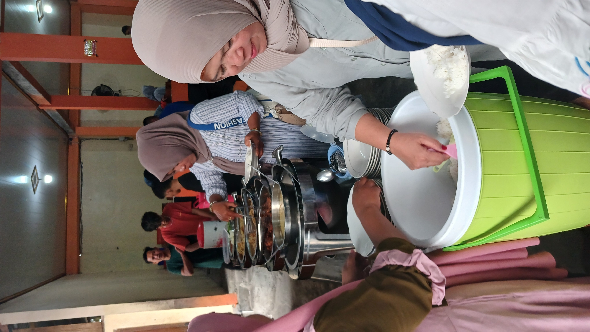 In Tanjungsari, Indonesia, a group of people are gathered around a buffet table laden with various food dishes. The scene appears to be an indoor setting, possibly a community hall or a restaurant, with orange-painted pillars and a beige-colored ceiling. The buffet is set up on a multi-tiered stainless steel stand, with chafing dishes holding food.
Several individuals are actively serving themselves or assisting others. On the right side of the frame, a woman wearing a light brown hijab and a grey shirt is holding a white plate and using a pink scoop to serve rice from a large green cylindrical container. Her gaze is directed downwards towards the rice. To her left, another woman in a pink hijab and a patterned top is holding a white plate, ready to be filled.
In the background, more people are visible, some standing and others in line. One person in a light brown hijab is using tongs to serve food from a chafing dish. A red and white cylindrical container is also visible behind the buffet table. The overall atmosphere suggests a mealtime event, likely a communal gathering or a catered meal.