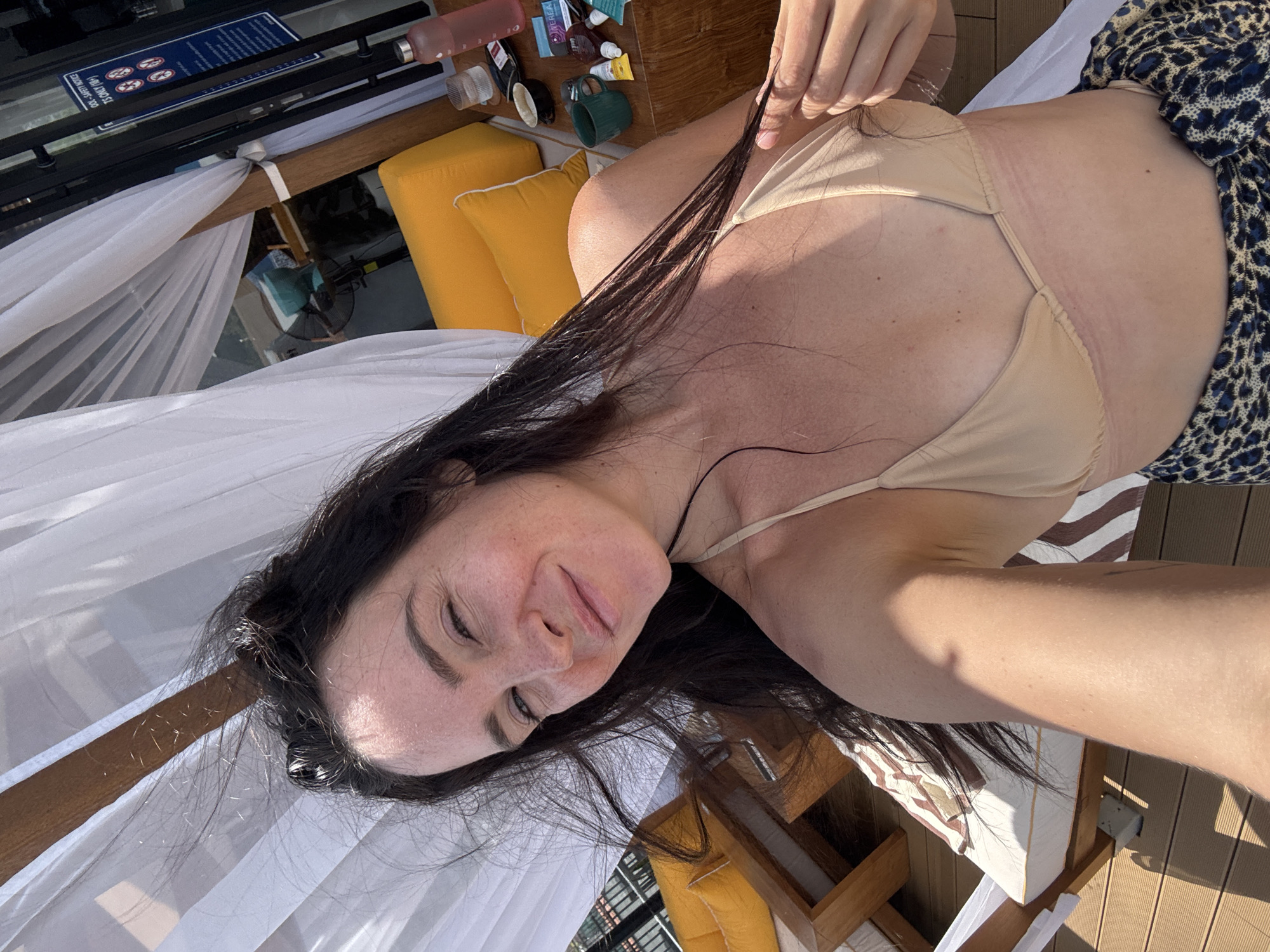A woman is taking a selfie outdoors, likely at a resort in North Kuta, Indonesia. She has long, dark hair that is partially windswept and she is wearing a tan bikini top. She is smiling and looking at the camera. Behind her, white curtains create a canopy, and yellow cushions and wooden furniture are visible, suggesting an outdoor seating area. A table with various items, including bottles and cups, is also in the background. The overall scene depicts a relaxed, sunny environment.