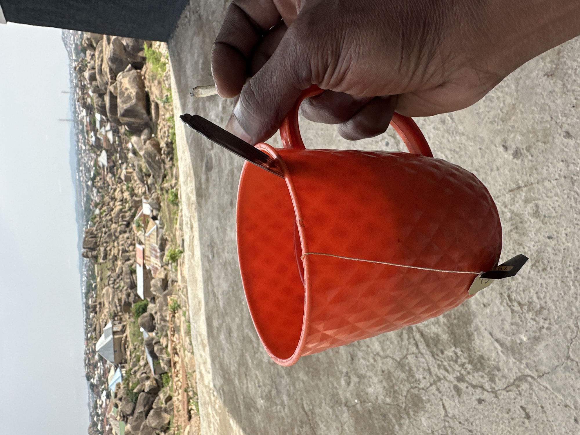 A man's hand holds an orange mug, a cigarette, and a black object that looks like a hair clip, outdoors. The backdrop is a cityscape of Jos, Nigeria, known for its rock formations and interspersed buildings. The concrete surface the objects are held over suggests a patio or balcony.