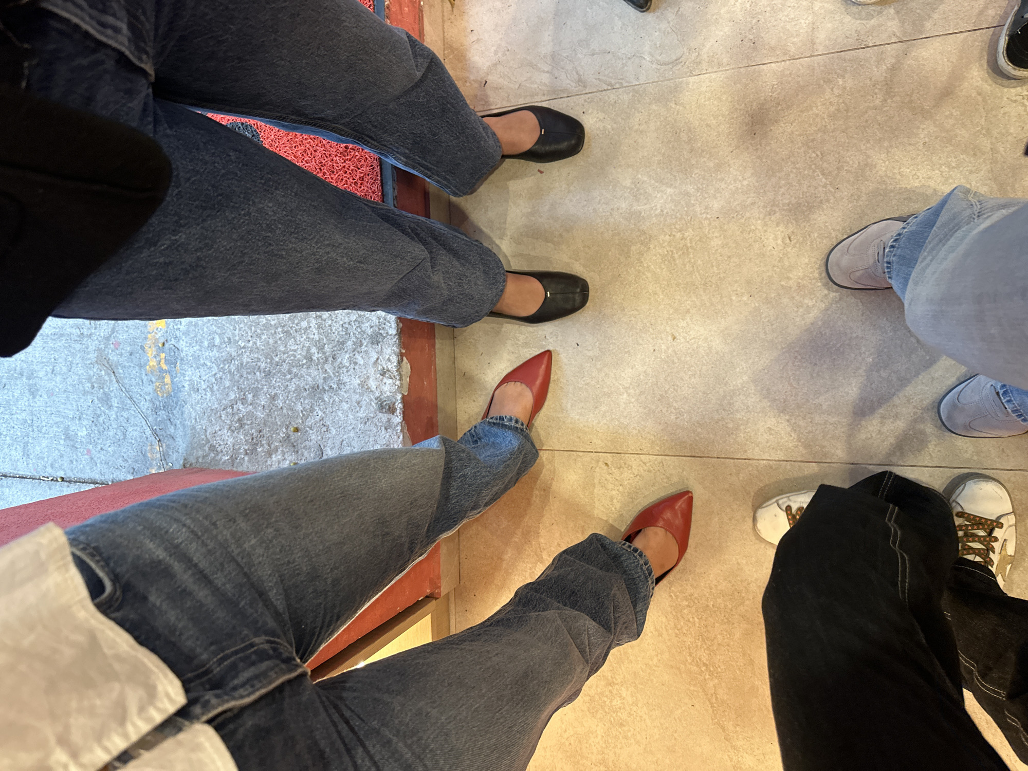 The image shows a close-up, overhead view of several people's lower bodies and feet, suggesting an indoor setting. From left to right, the first person wears dark wash jeans and black flats. The second person is wearing light wash jeans and pointed red heels. The third person is wearing light wash jeans and gray sneakers. The final person is wearing black jeans and white sneakers with gold stars and brown laces. The floor appears to be a light-colored, possibly tiled or concrete, surface. Part of a chair leg and red mat are visible on the left side. The image lacks context to determine the exact location or the nature of their interaction.