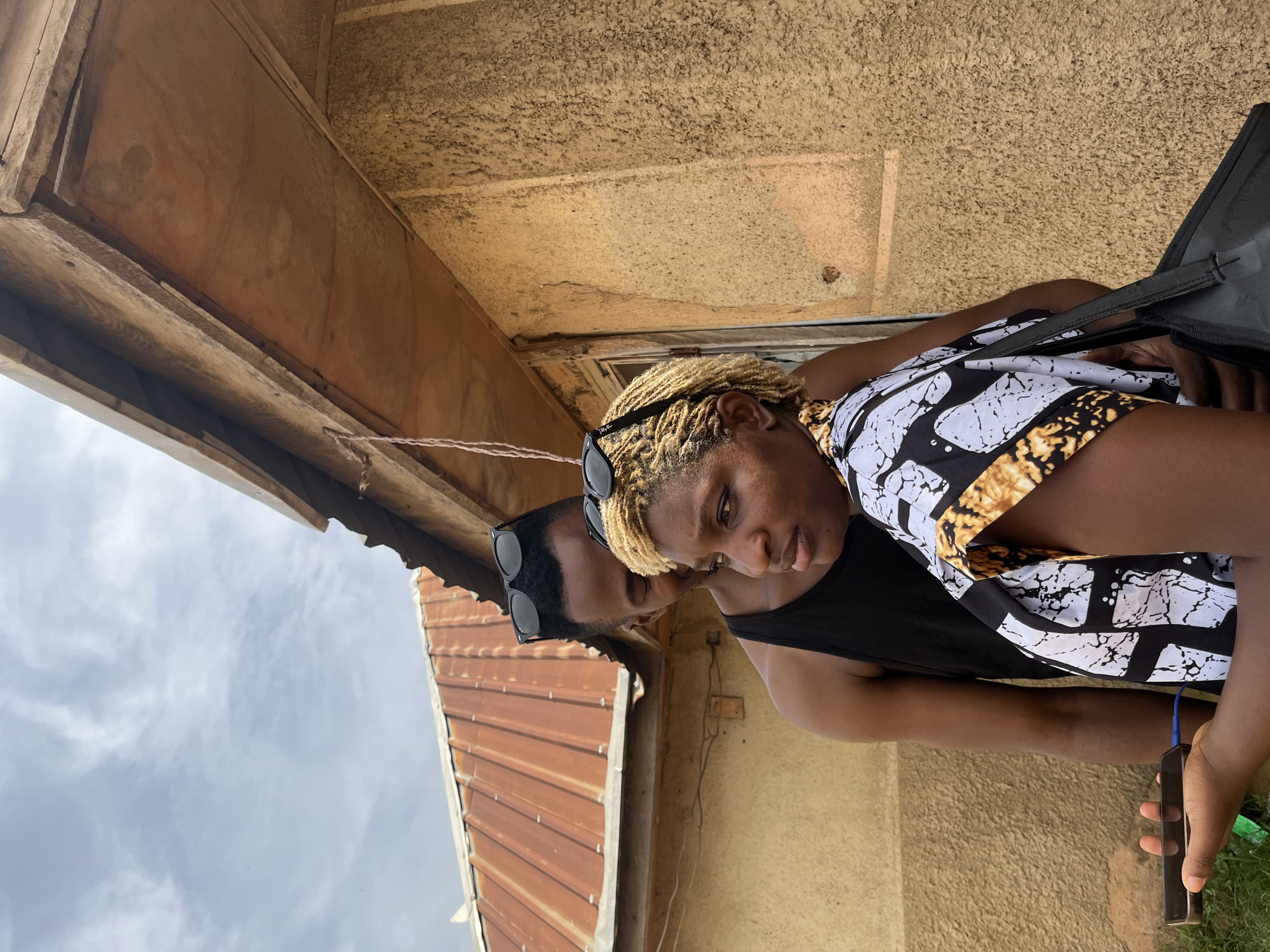 The image depicts two individuals, a man and a woman, standing outdoors in front of a textured, beige wall. The man, wearing a black tank top and sunglasses perched on his head, is positioned behind the woman, with his arms around her. The woman, facing to her left and looking downwards, has blonde, dreadlocked hair and is wearing a patterned black and white top with yellow accents. She also has sunglasses on her head. In her left hand, she holds a smartphone connected to a blue charging cable, with the charger itself obscured from view. A black bag is slung over her right shoulder. Above them, a corrugated metal roof, weathered and reddish-brown, is visible. The sky above is overcast with wispy clouds. The scene appears to be in a residential or semi-urban setting, potentially in the city of Jos, Nigeria, given the architectural style and vegetation, although this cannot be definitively confirmed from the image alone.
