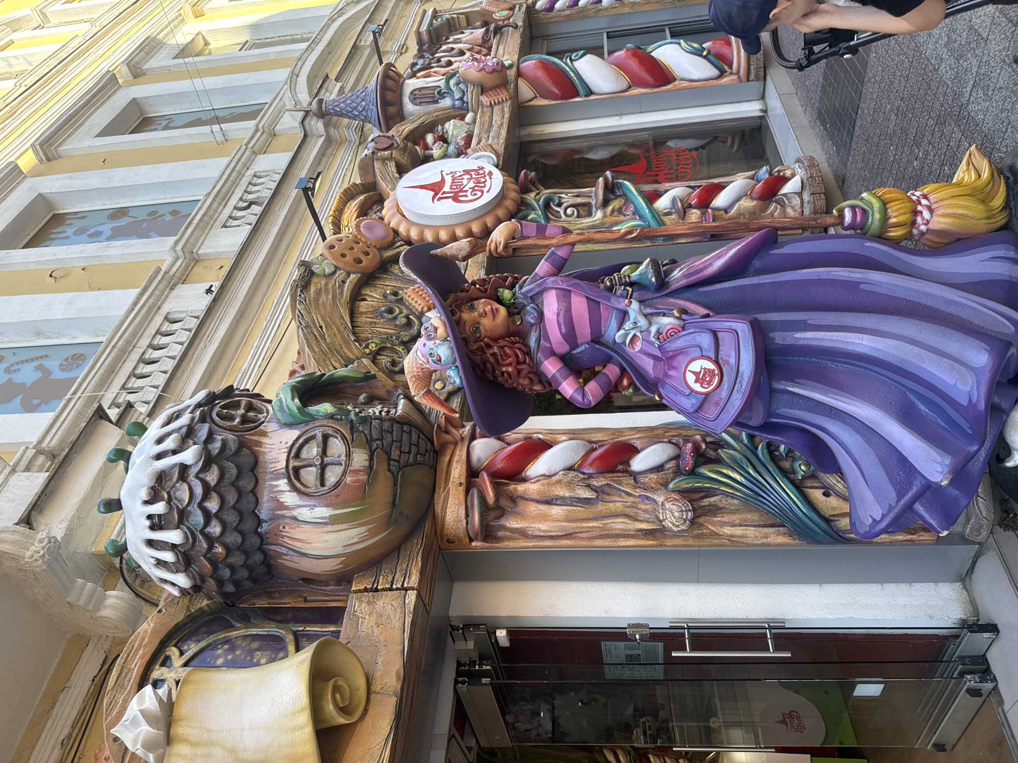 The image shows a whimsical, colorful facade of a building in Plovdiv, Bulgaria, likely a confectionery or candy store, adorned with fantastical elements. Dominating the foreground is a large, vibrant statue of a witch with curly red hair, a wide-brimmed purple hat, and a striped dress. She holds a broomstick and has a small mouse perched on her shoulder. The building's exterior is decorated with oversized, whimsical motifs resembling acorns, cookies, and other sweet treats, painted in warm earthy tones and bright accents. The entrance features a glass door with silver handles. Above the entrance, the building has windows with stylized silhouettes of candy. A sign with a red witch hat logo and the text "Hansel & Gretel" is visible. To the right, a young person wearing a dark cap is partially visible, looking towards the facade, seemingly with a stroller. The scene is set on a paved walkway, suggesting a public area.