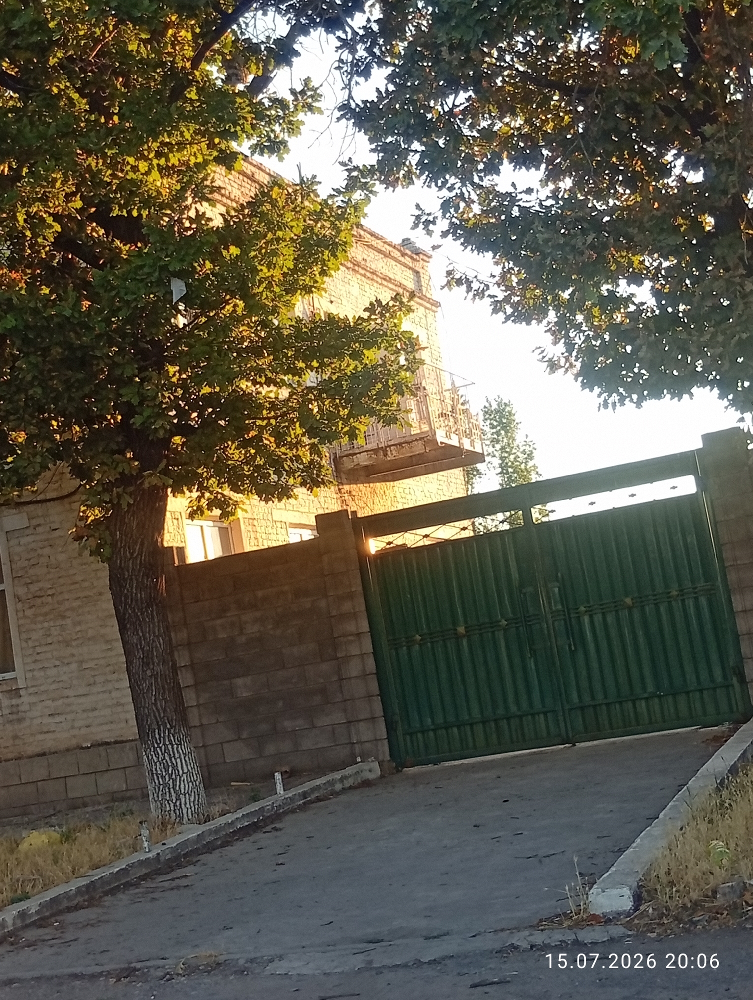 This is an eye-level, medium shot of a green, corrugated metal gate with two panels, set within a brick and stone wall. The gate is closed and appears to be made of metal. To the left of the gate, a large tree with a rough, grey-and-white patterned trunk and green leaves partially obscures a beige brick building. This building has windows with white frames and a balcony with a metal railing. Sunlight illuminates the scene, casting long shadows and highlighting the foliage. The ground in front of the gate is an asphalt driveway, bordered by a concrete curb and dry grass. A timestamp at the bottom right reads "15.07.2026 20:06". The location is identified as Ak-Suu, Kyrgyzstan. There are no people or animals visible in the image.
