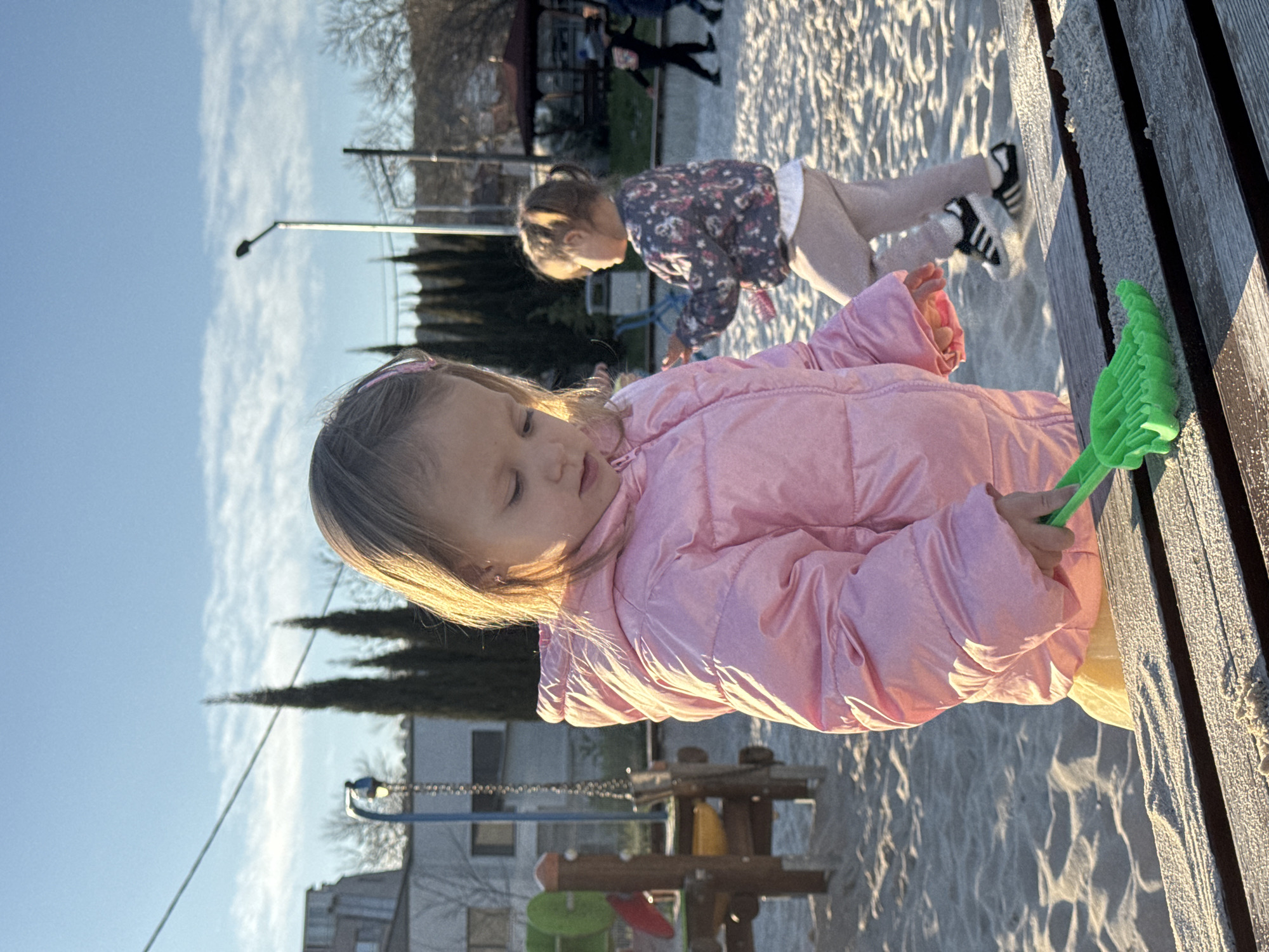 A close-up shot captures two young girls on a playground. The foreground girl, wearing a pink puffy jacket and a hairband, holds a green plastic rake. Behind her, another girl in a patterned shirt and light-colored pants kneels on the sand. Both are positioned near a wooden bench. The playground setting includes a building, swings, and lampposts. The location is likely Varna, Bulgaria, based on the architectural style of the building and general appearance. The image is taken during daytime, with clear sky.