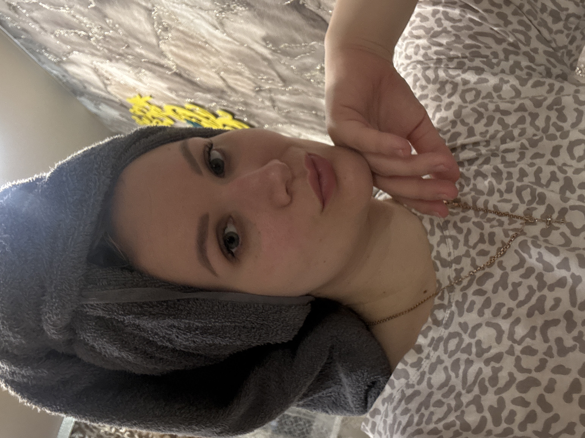 An indoor portrait features a young woman, shown from the chest up, with her head tilted and looking towards the camera. She has light skin, defined eyebrows, and light-colored eyes, with a neutral and relaxed expression. A dark grey, plush towel is wrapped around her head, suggesting a recent shower or a self-care moment. She is wearing a light-colored top with a distinct grey and off-white leopard print pattern. A thin, gold-colored chain necklace is visible around her neck. Her left hand is gently placed near her face, with fingers curled, resting against her jawline and cheek, adding to the relaxed pose. The background consists of a textured, light-colored surface, possibly a duvet or wall, with some blurred yellow elements towards the upper left. The lighting is soft and even, highlighting her features without harsh shadows. There is no visible text in the image. The scene conveys a sense of calm and domesticity in Chaikovsky, Russia.