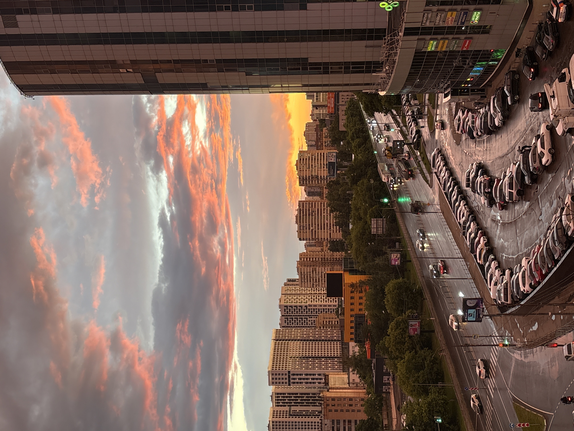 This media file captures a cityscape at dusk, with a dramatic sky filled with fiery orange and pink clouds. The scene is dominated by tall, modern buildings with glass facades, characteristic of an urban environment. In the foreground, a multi-lane road is visible, wet from recent rain, reflecting the streetlights and traffic signals. A row of parked cars, mostly white and dark-colored, lines the edge of the road. The sky transitions from bright oranges and pinks near the horizon to a softer grey, indicating the sun is setting.
Among the buildings, some display illuminated signage, suggesting they are commercial establishments or offices. The overall atmosphere is one of urban activity winding down as night approaches. The wet pavement and overcast sky hint at recent or ongoing precipitation. There is no clear indication of people in the frame, but the presence of cars and buildings points to a populated area. The time of day is clearly dusk, with the dramatic cloud colors being a key feature. The location is not specified but appears to be a modern city. A few signs with text are visible on the buildings, but they are not legible enough to decipher specific words.
