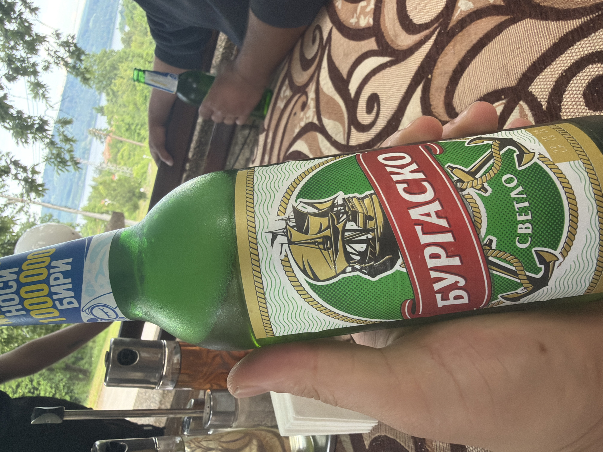 The image, taken in Sushina, Bulgaria, captures a serene outdoor scene centered around a person holding a green beer bottle. The foreground is dominated by a hand gripping a bottle of "Burgasko" beer. Its label features a sailing ship, anchors, and rope details, with prominent Cyrillic text reading "БУРГАСКО" (Burgasko) and "СВЕТЛО" (Svetlo), indicating a light beer. A blue and white neck label further states "НОСИ 100 000 000 БИРИ" (Nosi 100,000,000 Biri), a promotional slogan, along with small numbers "12 10 23" possibly indicating a date.
In the mid-ground, part of another person, seated at a table, is visible, holding a second green bottle. The table is covered with a patterned brown and cream tablecloth, and metal condiment dispensers are also partially visible.
The background reveals a picturesque daytime landscape from a high vantage point. A vast, calm body of water, likely a river or lake, stretches across the mid-distance, surrounded by lush green, tree-covered hills or mountains under a clear sky. The setting suggests a relaxing moment, possibly at an outdoor cafe or restaurant, enjoying drinks with a scenic view in good weather.