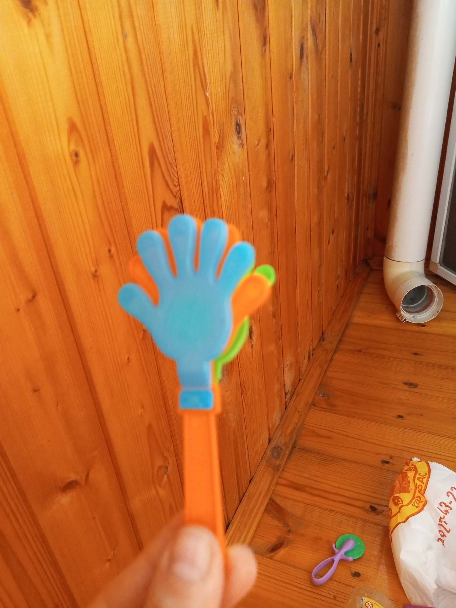 An out-of-focus close-up shot shows a person's hand holding a colorful plastic toy shaped like a clapping hand. The toy is orange with a light blue hand and a green accent. The background consists of light brown wooden paneling on the wall and floor. To the right, a white pipe extends upwards, and a white plastic bag with red lettering is visible on the floor. The scene appears to be indoors, likely in a home. The image does not provide enough information to determine the specific location within the city of Chistopol, Russia.