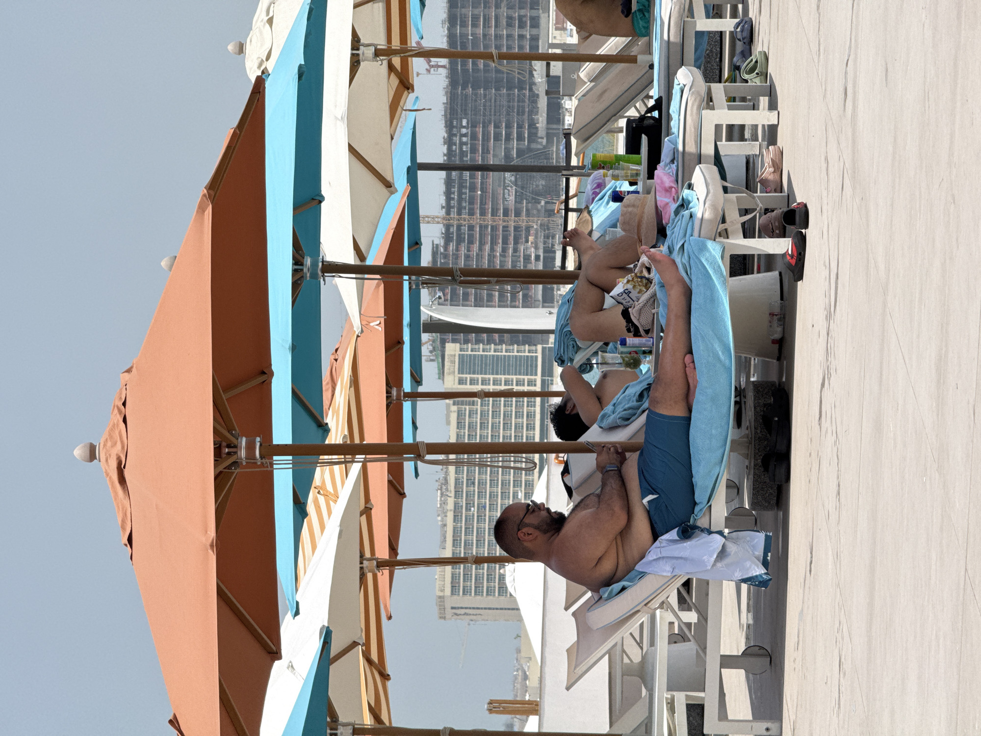 This image depicts a poolside scene in the United Arab Emirates, possibly Dubai, given the architectural style of the buildings in the background which are under construction. The foreground features two individuals relaxing on lounge chairs under large umbrellas.
The person on the left, a man with glasses and a shaved head, is wearing blue swim trunks and appears to be using his mobile phone. He is reclining on a white lounge chair covered with a blue towel and a white garment draped over its side. To his right, another person is also reclining, partially visible, with their head resting on a towel.
Several lounge chairs are arranged on a light-colored tiled surface. A variety of beach accessories are scattered around, including towels, a hat, sunglasses, bottles of beverages, and a bag. Multiple large umbrellas, some with orange and turquoise canopies, provide shade. In the background, several multi-story buildings are visible, some of which are clearly under construction, with cranes and scaffolding indicating ongoing development. The sky is bright and clear, suggesting a sunny day.