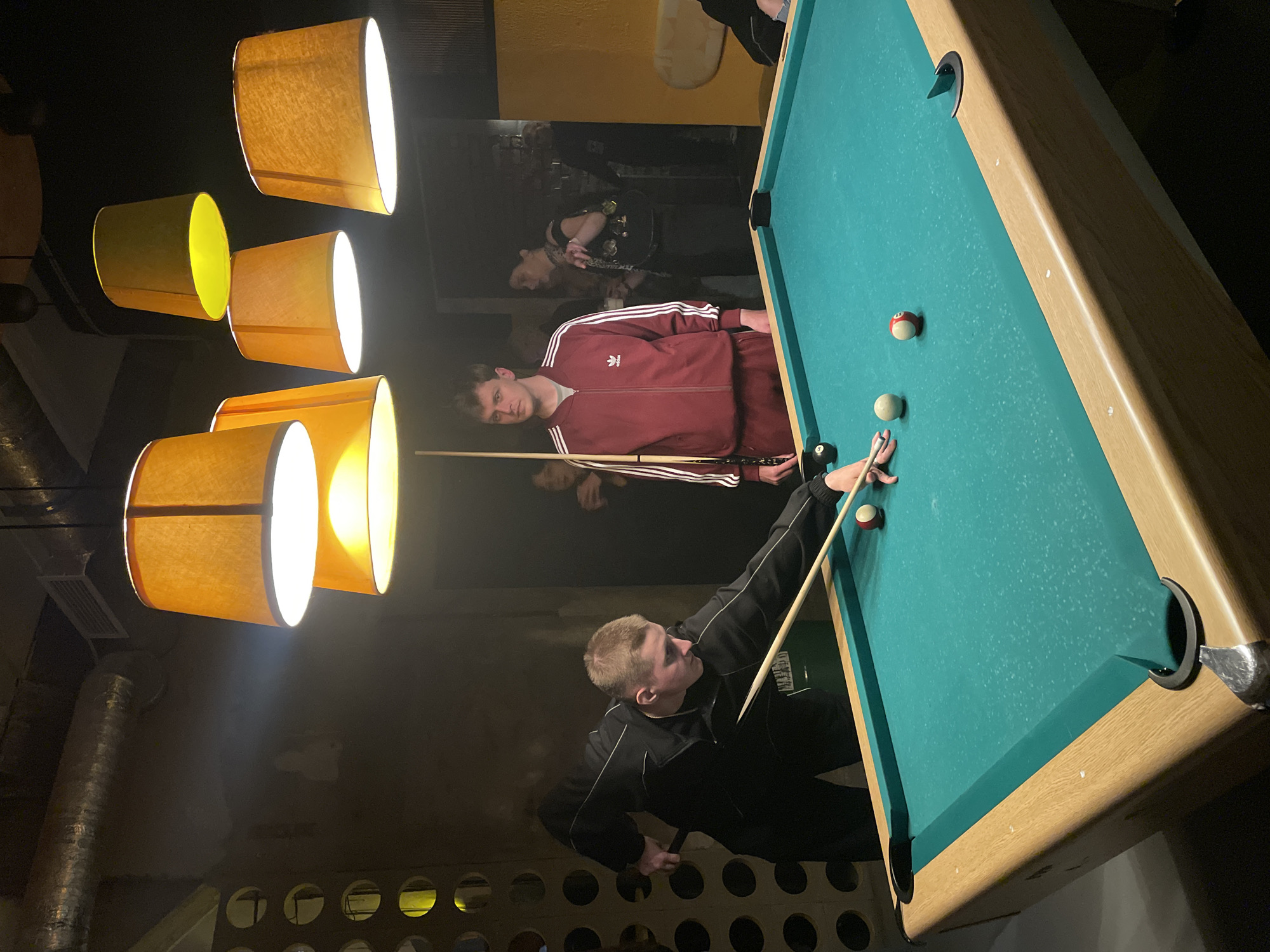 The image captures a scene inside a dimly lit establishment, likely a bar or recreation area. Two individuals are positioned around a pool table, which is the central focus. One person, dressed in a black tracksuit, is in the process of taking a shot, cue in hand, with a focused expression. Another individual, wearing a red Adidas tracksuit, stands and watches. The pool table is green with multiple balls visible.
In the background, other people can be seen, though they are less sharply focused. Above the pool table, a cluster of several lamp shades hang, casting an ambient yellow light. The walls show signs of wear and character, with a visible design of yellow circular shapes on one side. Based on the interior architecture and ambiance, this could be a scene from a bar or recreational center, potentially in Saint Petersburg, Russia.