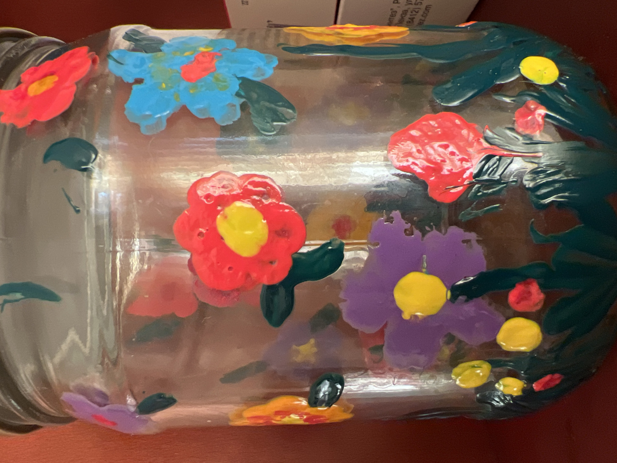 The image shows a close-up of a clear glass jar adorned with colorful, hand-painted flowers. The flowers are depicted in various vibrant colors including red, yellow, purple, blue, and orange, with some featuring a yellow center. The paint appears thick and slightly raised, giving a textured appearance. Green stems and leaves are also visible, creating a garden scene on the jar's surface. The jar has a metallic lid. The background is out of focus, showing a reddish-brown surface and a portion of a cardboard box with some text, including what appears to be a phone number. The lighting suggests an indoor setting. There are no people present in the image. The image does not contain any information about the location in Minsk, Belarus.