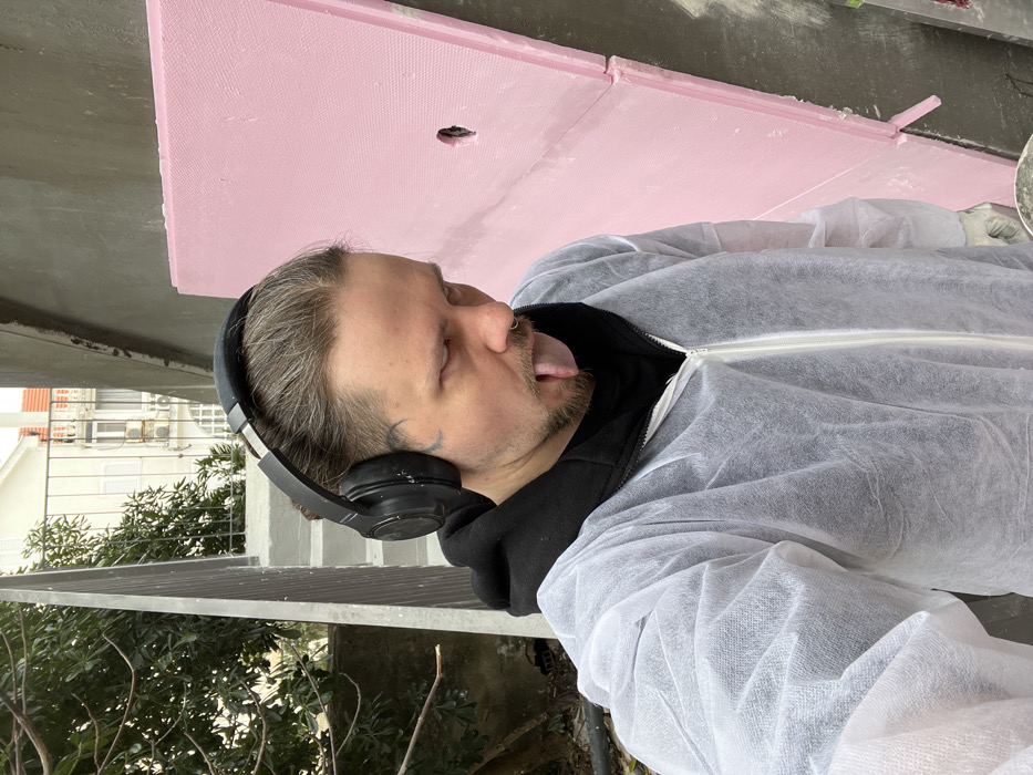 An adult male is prominently featured in a close-up outdoor scene in Bogdašići, Montenegro. He is positioned slightly off-center, facing the camera with his tongue extended. The individual has dark blonde or light brown hair tied back, a beard, a nose ring on his left nostril, and a dark tattoo or marking on his right temple/upper cheek area. He is wearing a black hoodie under a white disposable protective coverall with a zippered front, and black over-ear headphones are on his head. To the subject's right (viewer's left), a textured, light gray wall, possibly concrete or plaster, is visible. To the subject's left (viewer's right), a large, rectangular pink insulation board with a visible circular cutout is affixed vertically against a wall. The lower right portion of the image shows a glimpse of a gloved hand holding a small container with white material. In the background, beyond a gray metal railing, green foliage partially obscures a light-colored building featuring windows, balconies, and an external air conditioning unit under an overcast sky. The overall scene suggests ongoing construction or renovation activity.