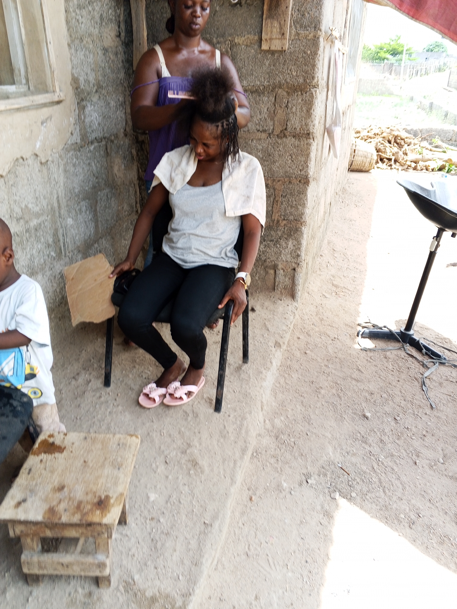 Outside a building in Adonkolo, Nigeria, a woman in a purple tank top is braiding the hair of a seated woman wearing a gray shirt, black pants, and pink sandals. A young boy in a white shirt stands nearby. A small, wooden stool is in the foreground. A microphone stand sits on the ground to the right. Behind the building is a pile of wood debris.