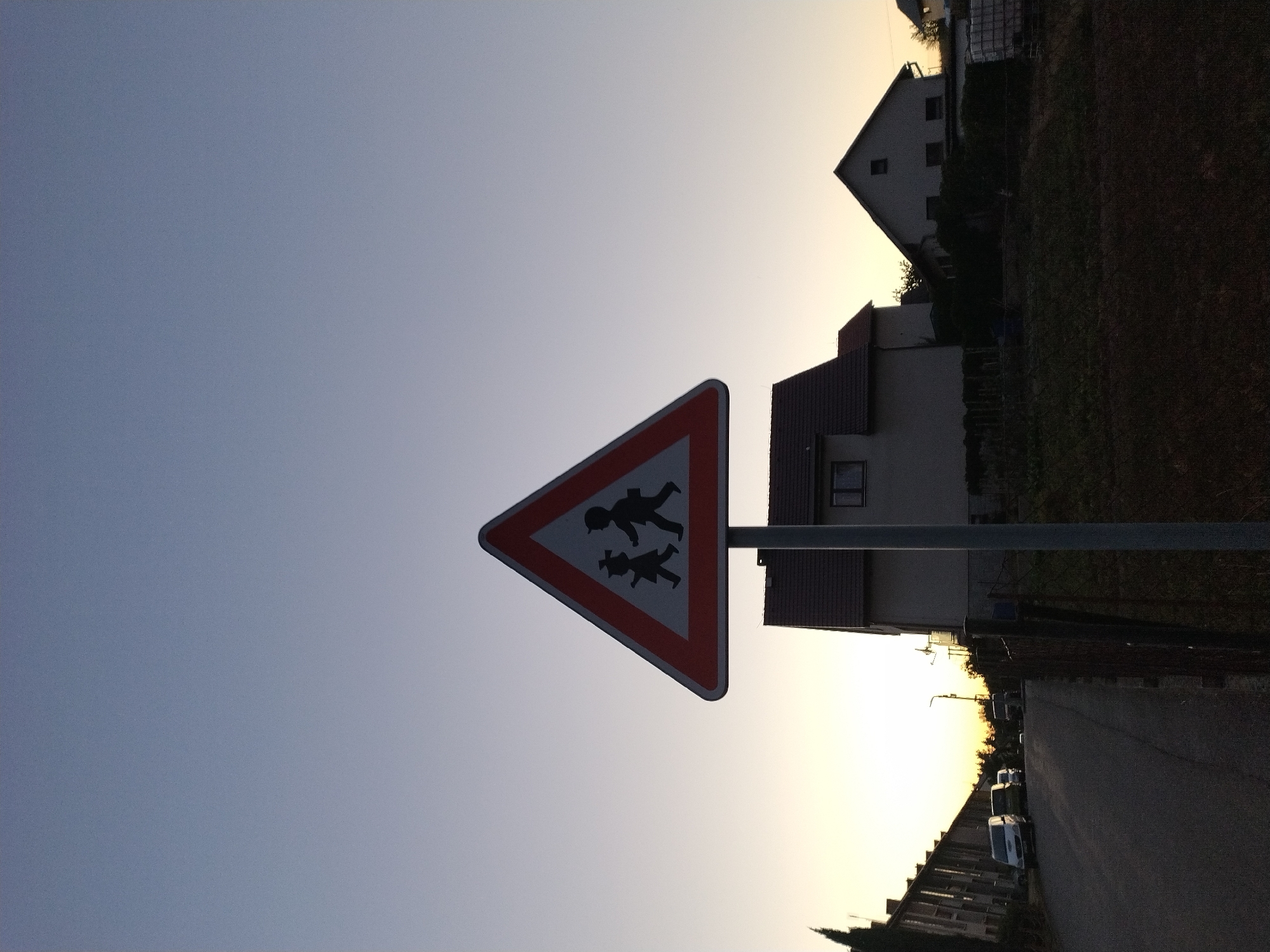 A triangular road sign warning of children crossing is centrally located in the foreground, silhouetted against a clear, pale blue sky that transitions to a soft yellow at the horizon. The sign features a red border and depicts two black stick figures, one appearing to be a child wearing a hat, walking from left to right. Behind the sign, a rural residential area is visible at dusk. To the left, a street is lined with parked cars, including a white van. Further back, several houses with pitched roofs are discernible, with one prominent house featuring a visible window. The environment is characterized by the fading light of sunset, casting a serene atmosphere over the quiet street and suburban houses. The image does not contain any visible people or specific actions occurring.