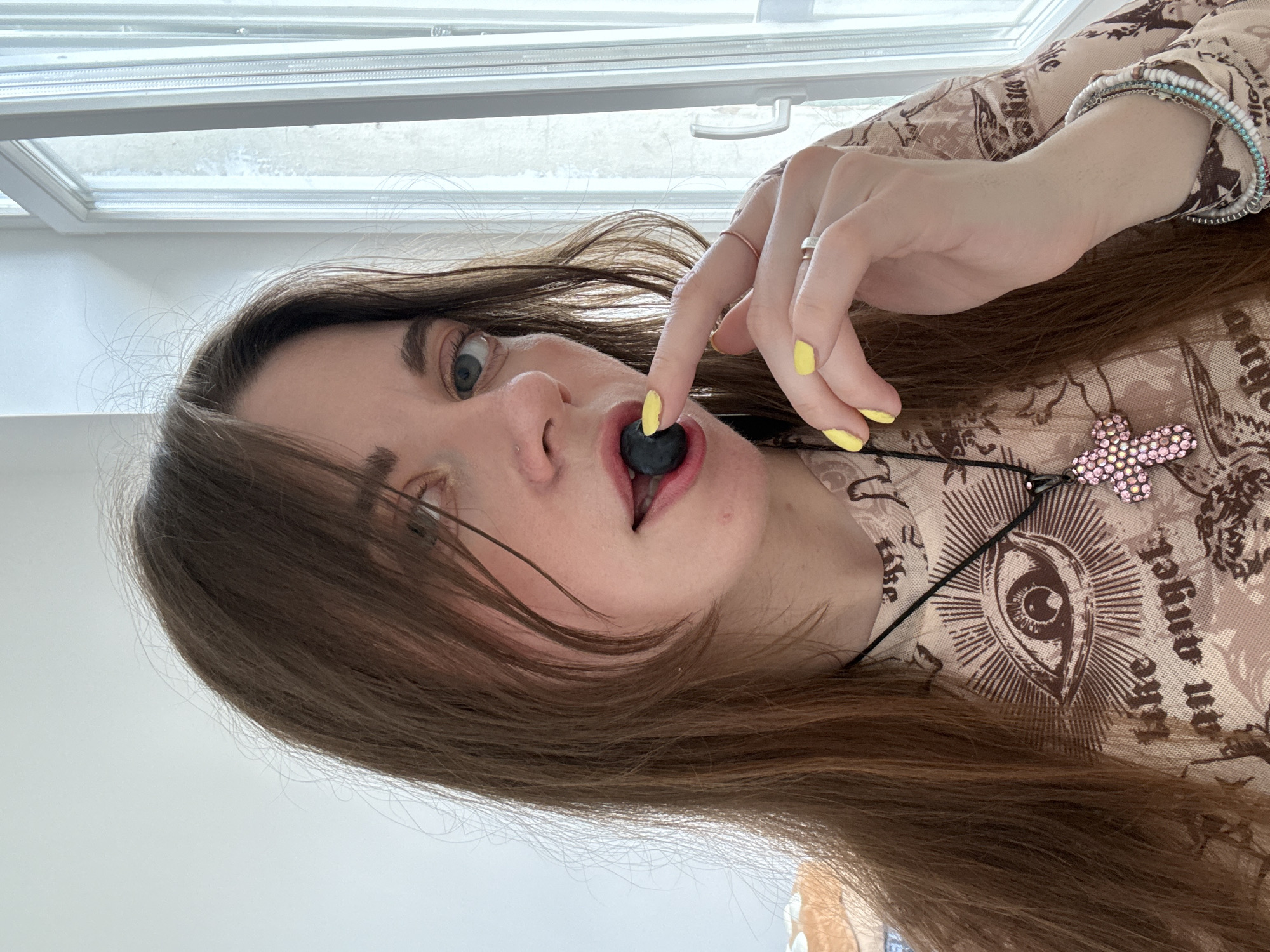 A woman is holding a blueberry in her mouth. Her nails are painted yellow. She is wearing a patterned top and a necklace with a pink butterfly pendant. She has bracelets on her wrist and a ring on her finger. The background shows a window. The image does not contain enough information to determine a location in Moscow, Russia.