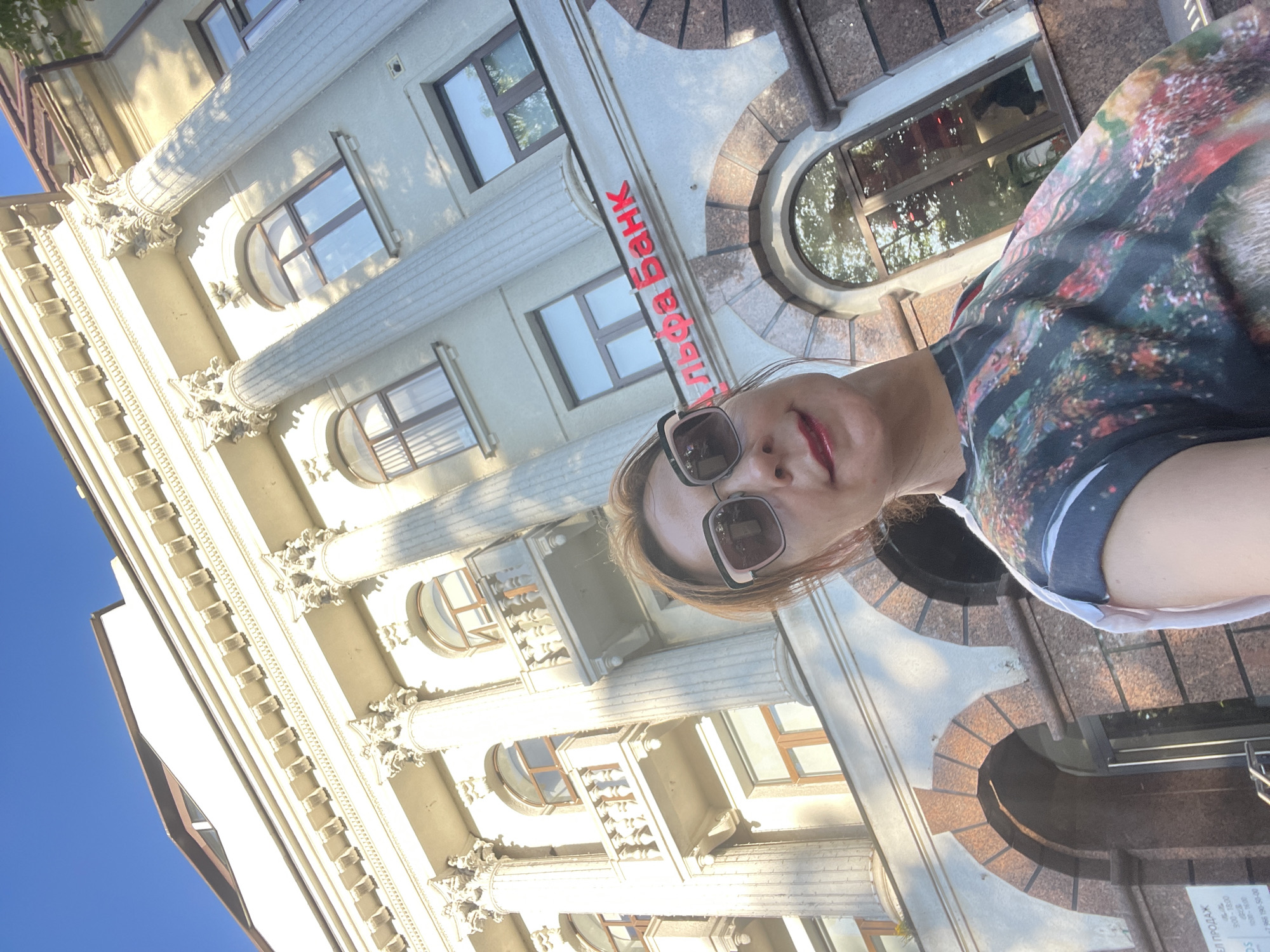 A woman with auburn hair and dark sunglasses takes a selfie outdoors, with a large, classical-style building in the background. The building has columns, balconies, and arched windows. Red lettering on the building reads "Альфа Банк," identifying it as an Alfa-Bank branch. The woman is wearing a colorful, patterned t-shirt. The sky above is clear and blue.