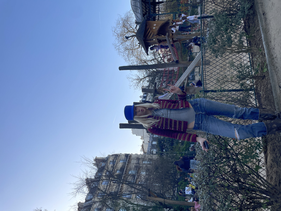 A blonde female individual is positioned centrally in the foreground of a public park in Paris, France, under a clear blue sky. She wears a blue cylindrical hat, a cropped maroon jacket with gold horizontal stripes, a light grey bodysuit or top, ripped blue jeans, and dark boots. Her right hand displays a "rock on" gesture, while her left hand holds a dark-colored smartphone. Her mouth is slightly open with her tongue visible, and her eyes are directed forward. Behind her, a low line of green bushes and a dark metal fence separate the immediate foreground from a busy playground area.
In the mid-ground to background, a multi-level wooden playground structure with a slide and red climbing nets is actively occupied by several children. Other adults and children are dispersed throughout the park, some near the playground and others in the distance. Deciduous trees with bare branches are visible across the scene, indicating a cold season. In the far background, typical Parisian Haussmannian apartment buildings with mansard roofs and multiple windows line the street. A faint white contrail is visible in the upper right portion of the clear blue sky.