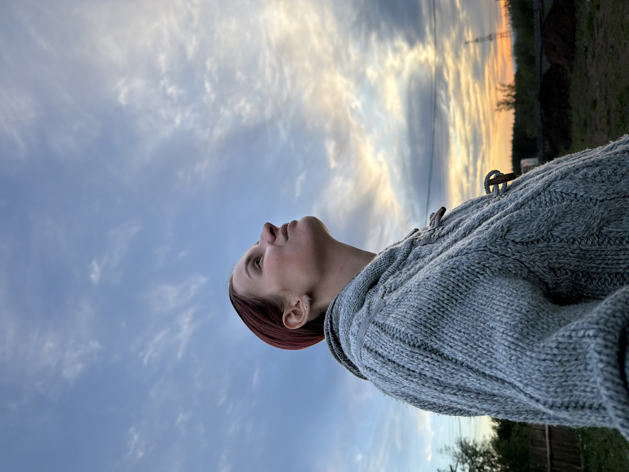 A young woman with reddish-brown hair looks up at the sky. She is wearing a gray cable-knit sweater. The sky is blue with white and golden clouds. Power lines and trees are visible in the background. It is dusk. The image appears to be taken outdoors in the vicinity of the city of Chaikovsky, Russia, judging by the visual cues of the environment and the prompt's explicit reference.