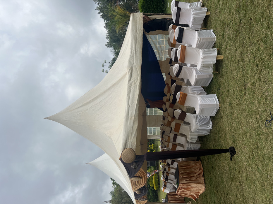 This image captures an outdoor event setup on a well-maintained grassy lawn in Makuyu, Kenya, under an overcast sky. The primary subjects are two large, light-colored pyramid-shaped tents, or marquees, with distinct pointed tops. The larger tent, positioned prominently, features a blue fabric lining or drape visible inside.
Beneath the main tent, numerous chairs are neatly arranged in rows, suggesting seating for a gathering. Each chair is covered in white fabric and adorned with sashes or ribbons in alternating colors of brown, black, and a golden or orange shade. Several tables are also visible, draped with gold or brown table skirts. Decorative elements, including woven, circular objects resembling wicker and dark fabric drapes, are strategically placed along the tent's supports. A person is partially visible near the top of the larger tent, appearing to be involved in its setup or adjustment.
The setting is a natural outdoor environment, characterized by expansive green grass and lush trees and foliage in the background. The sky is uniformly gray and cloudy, indicating an overcast day with diffused lighting. The overall scene suggests that an organized event, possibly a wedding, celebration, or formal gathering, is either being set up or is ready to commence. No discernible text is visible in the image.