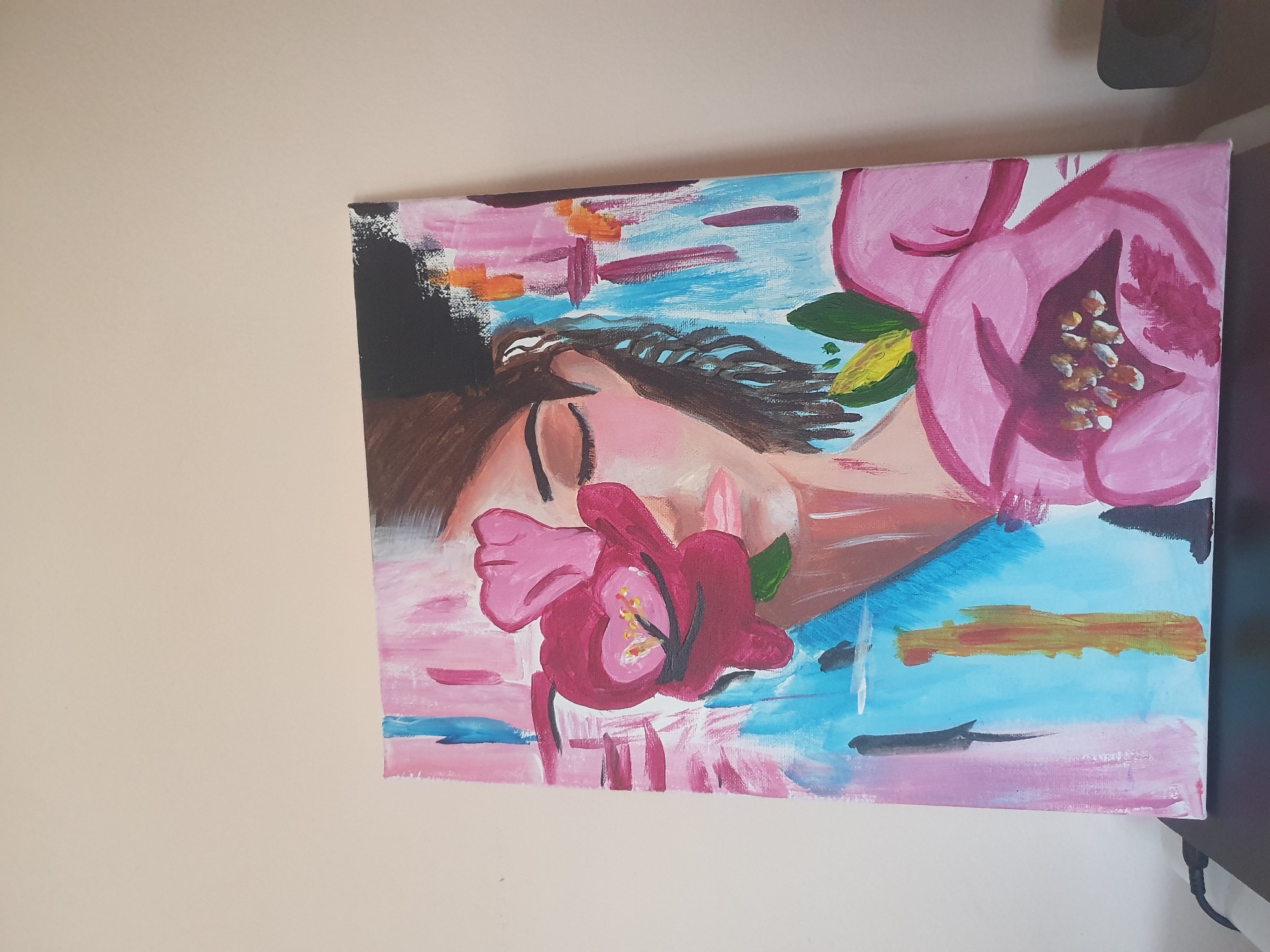 The image is a close-up of a rectangular canvas painting. The painting depicts a person lying down amidst flowers. The person has dark brown hair at the top left of the canvas, transitioning into dark curly hair across their forehead. The person's skin is a pale pinkish tone. The painting features pink flowers with green leaves. The background is painted with a mixture of pink and blue brushstrokes. The painting is hanging on a pale yellow wall. No location in Plovdiv, Bulgaria is evident.