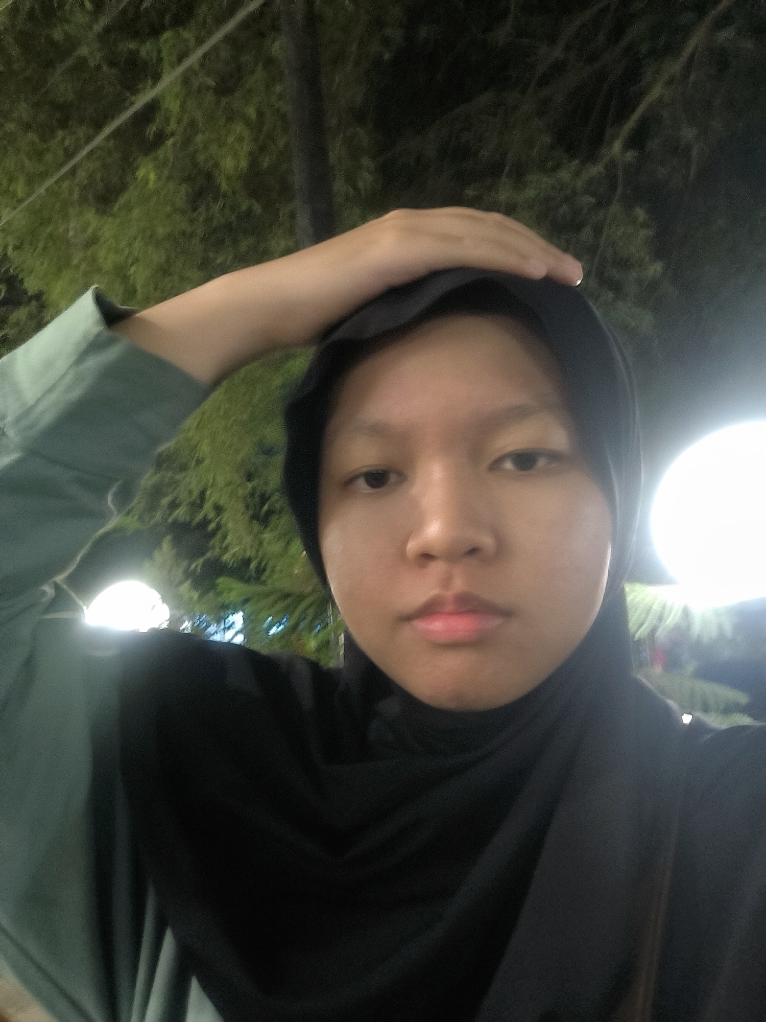 A selfie taken at night shows a young person wearing a black hijab and a green long-sleeved shirt. Their right hand is raised to touch the top of their head. The background features out-of-focus trees and bright, circular lights, suggesting an outdoor setting in a city like Surakarta, Indonesia, possibly at an event or a park illuminated for evening. The lighting indicates it is after sunset.