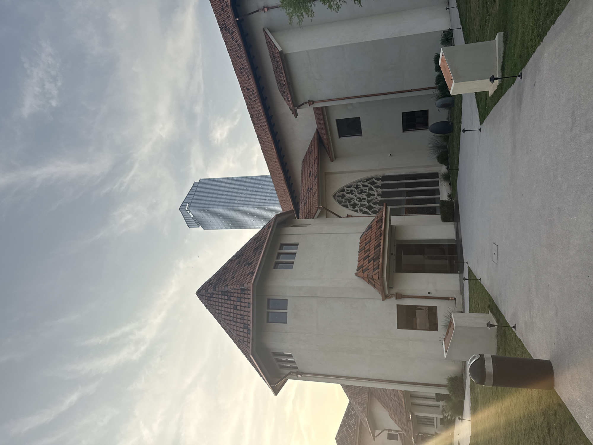 The image depicts a building with a red tile roof and white walls in San Pedro Garza García, Mexico. A modern skyscraper stands in the background, juxtaposed with the older architectural style of the building. A concrete path runs along the side of the building with lamp posts and green landscaping separating the building and path. A trash receptacle is visible in the foreground. The sky is cloudy.