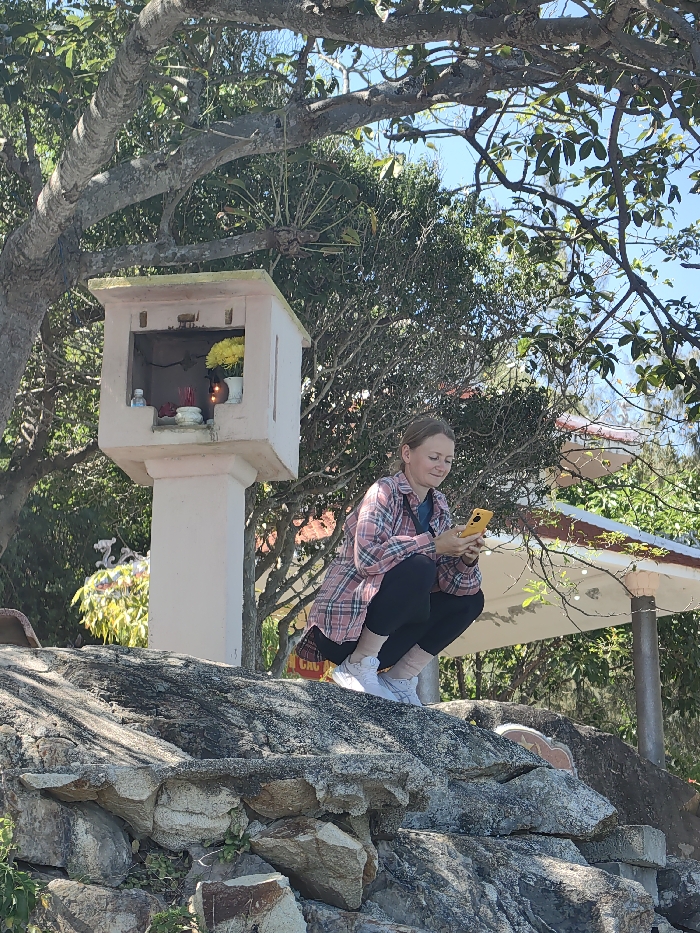A woman crouches on a rock, engrossed in her yellow phone. She wears a pink plaid shirt over black leggings and white sneakers. Behind her stands a small white shrine adorned with flowers and incense. The background includes lush green trees, a clear blue sky, and a glimpse of a white pavilion with a red-tiled roof. The scene is bathed in bright sunlight.