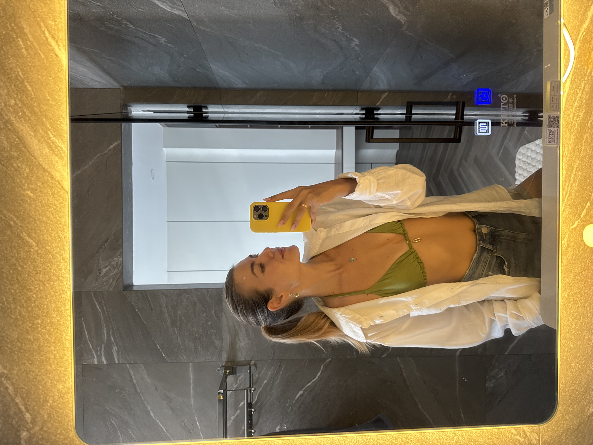 A person is taking a selfie in a bathroom mirror. The person is wearing a green bikini top, denim shorts, and a white button-down shirt. They are holding a yellow smartphone and facing to the left. The background includes gray marble walls, a white cabinet, and a illuminated mirror. There is a towel rack on the wall. The scene is taking place in an interior location.