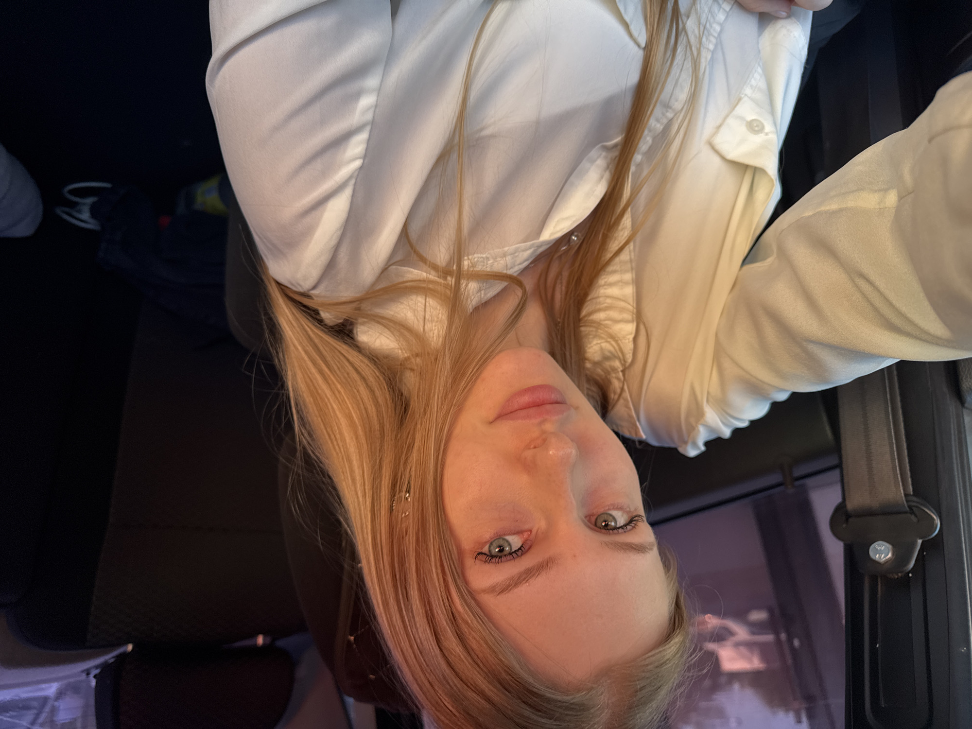 A young woman with light blonde hair is in a vehicle, presumably a car. She is wearing a white collared shirt. The background is blurry but shows a window with a partially visible street scene. A black seatbelt mechanism is visible to her right. While it isn't possible to determine her specific location in Perm, Russia, the scene suggests a car interior with an outside view through the window.